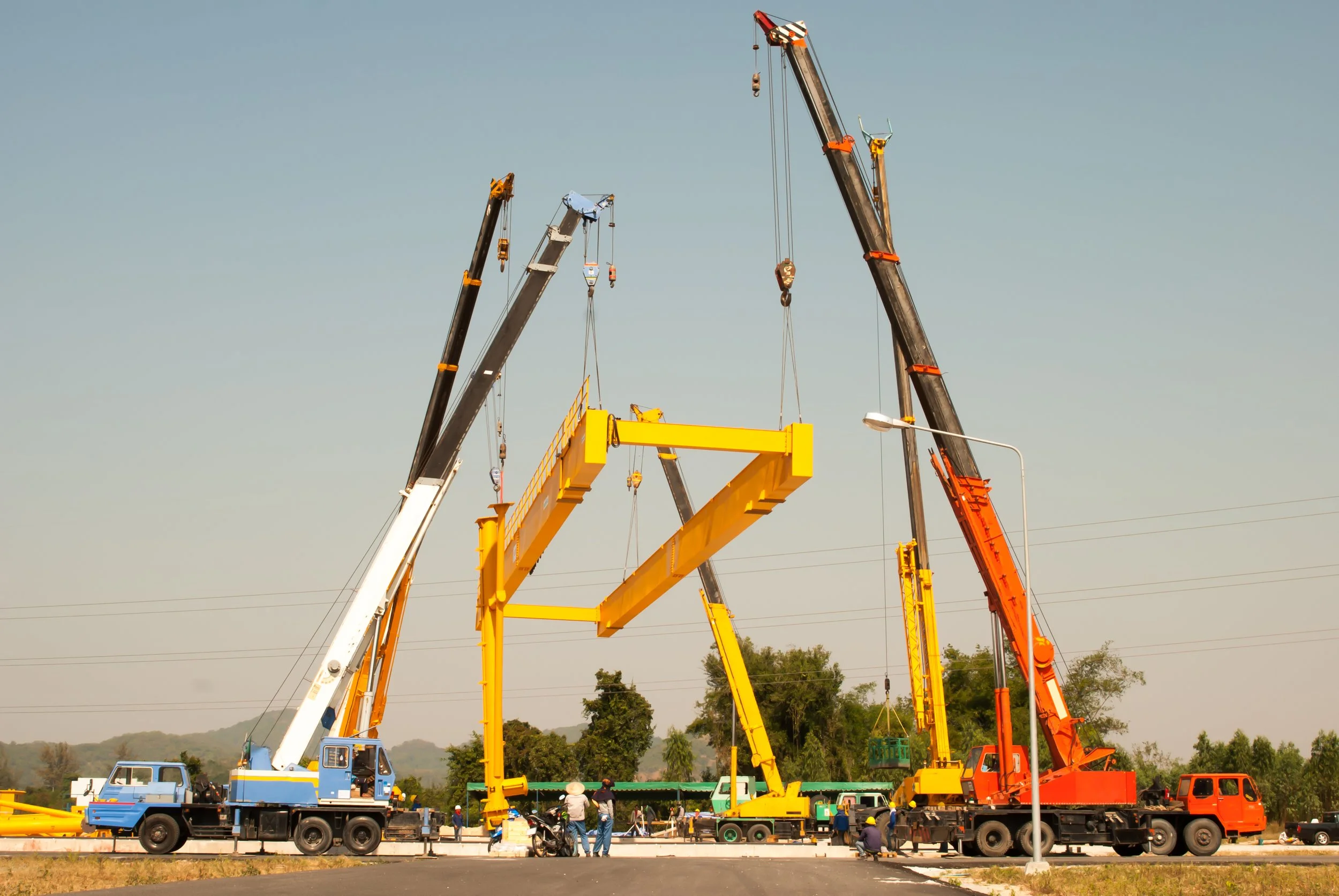 Construction site with multiple large cranes lifting yellow steel structures, workers and vehicles nearby, set against a clear sky and trees.
