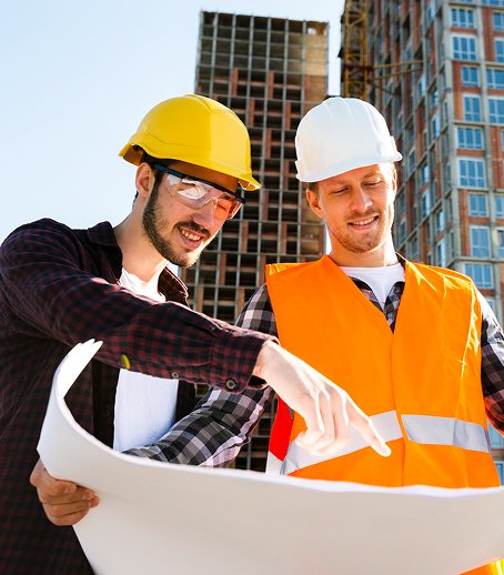 Two construction workers wearing safety helmets and reflective vests reviewing blueprints at a construction site with buildings in the background.