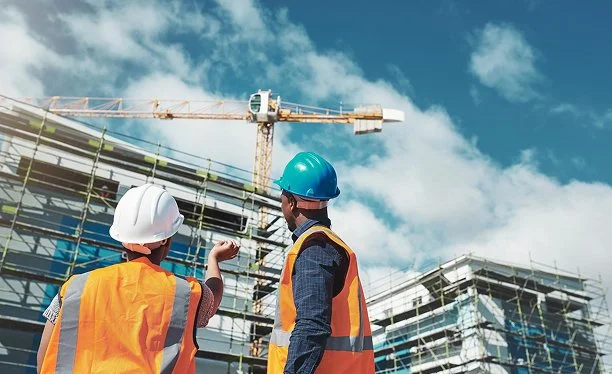 Two construction workers wearing safety vests and helmets standing on a construction site with scaffolding and a crane.