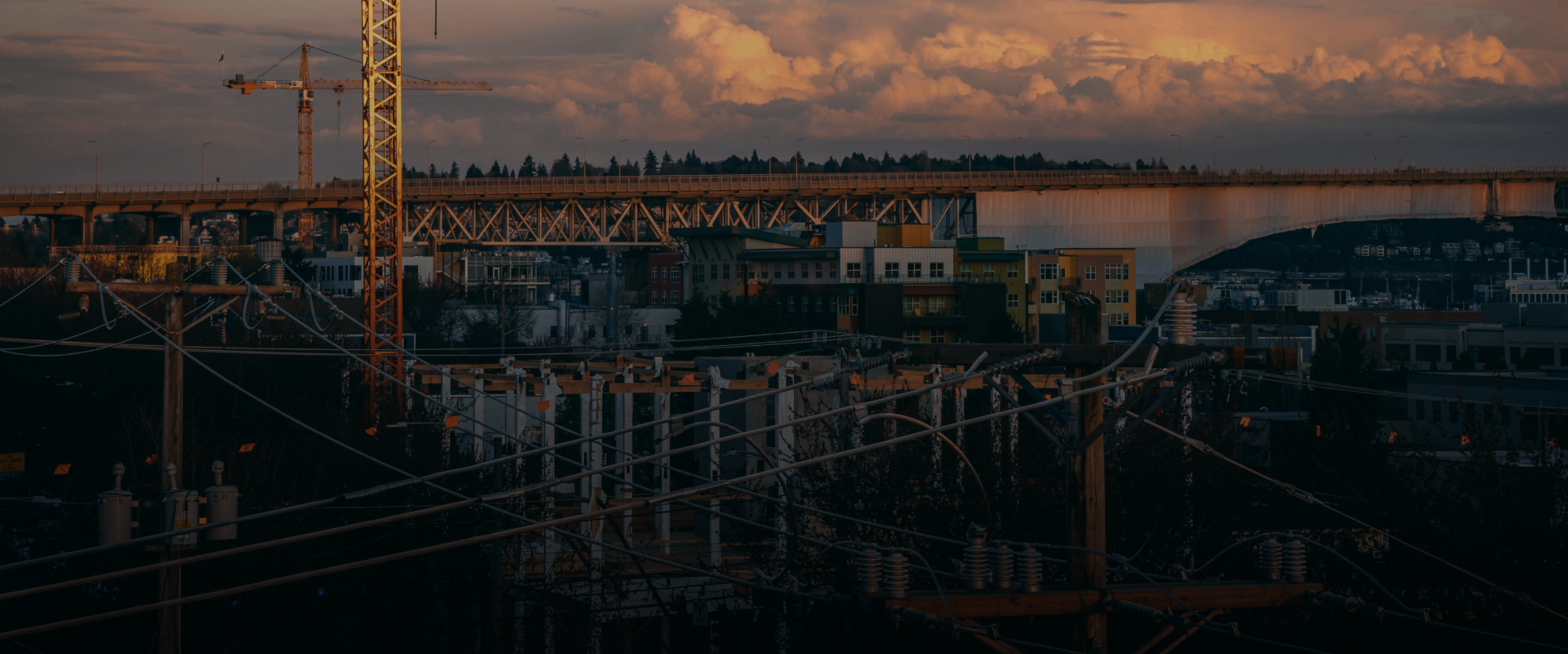 An urban landscape at sunset with a large bridge, construction cranes, power lines, and buildings under a cloudy sky.