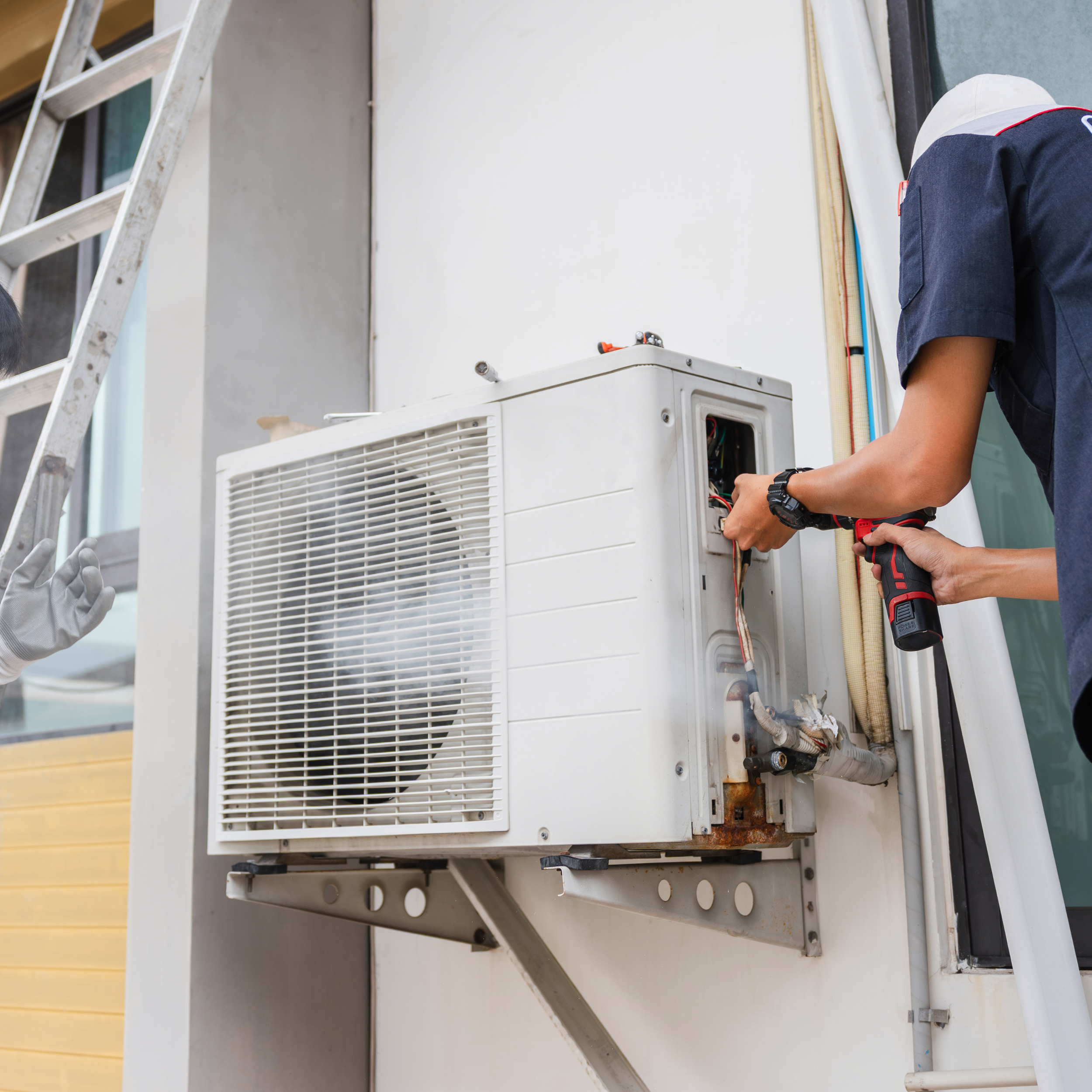 A person with a white cap and dark blue uniform working on an outdoor air conditioning unit using a cordless drill, with a ladder and colored pipes visible in the background.