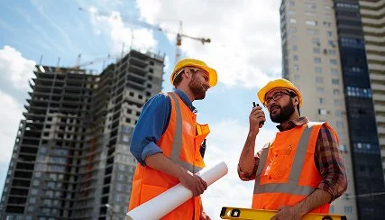 Two construction workers in yellow helmets and orange safety vests having a discussion on a construction site with tall buildings in the background.