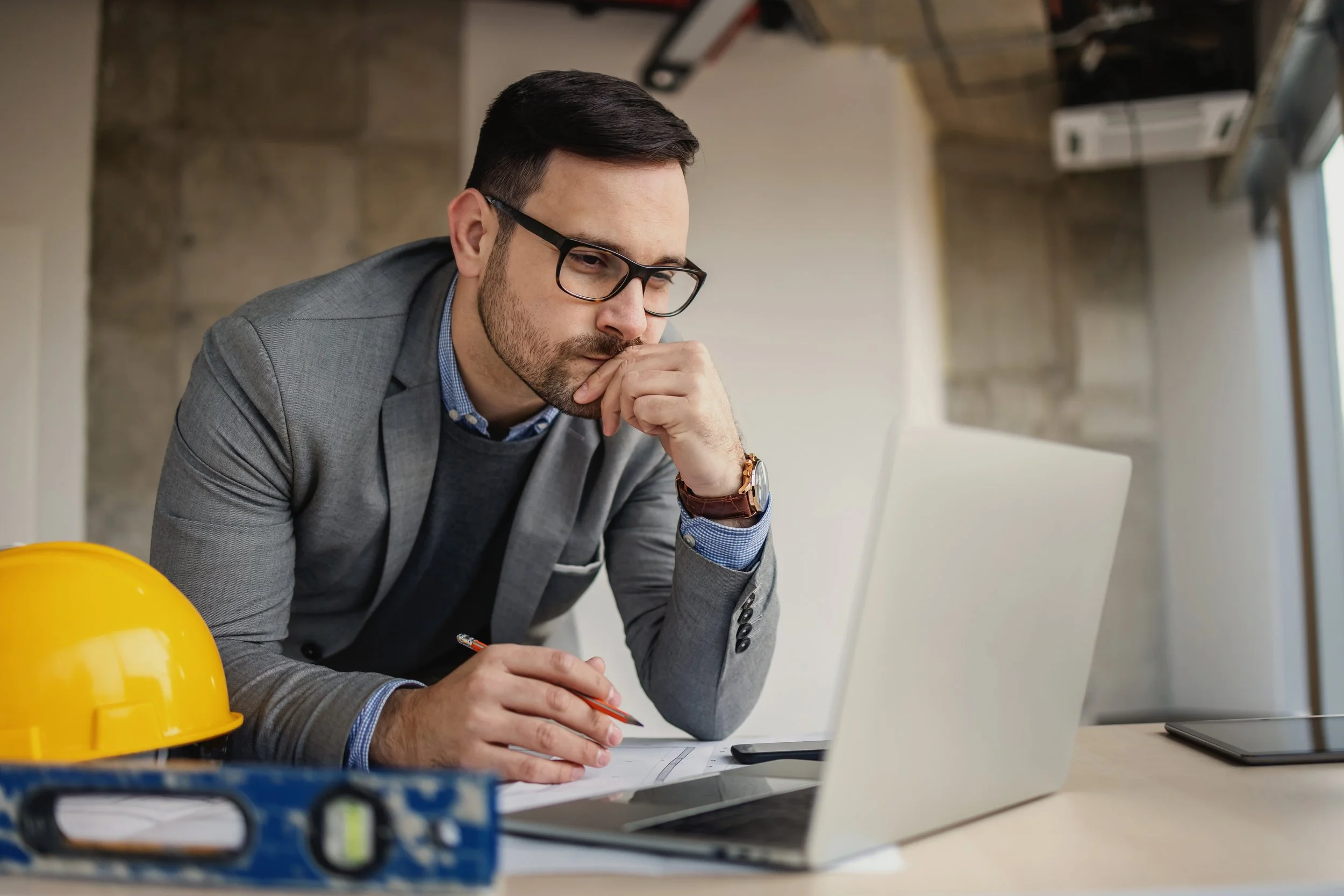 Man in a gray blazer and glasses working at a desk, surrounded by construction equipment like a yellow hard hat and blueprints, with a laptop open in front of him.