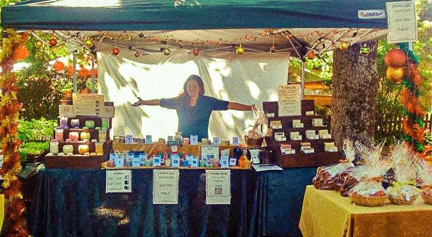 A woman standing behind a booth at an outdoor market, displaying various products like soaps and lotions, with festive decorations and colorful ornaments hanging around.