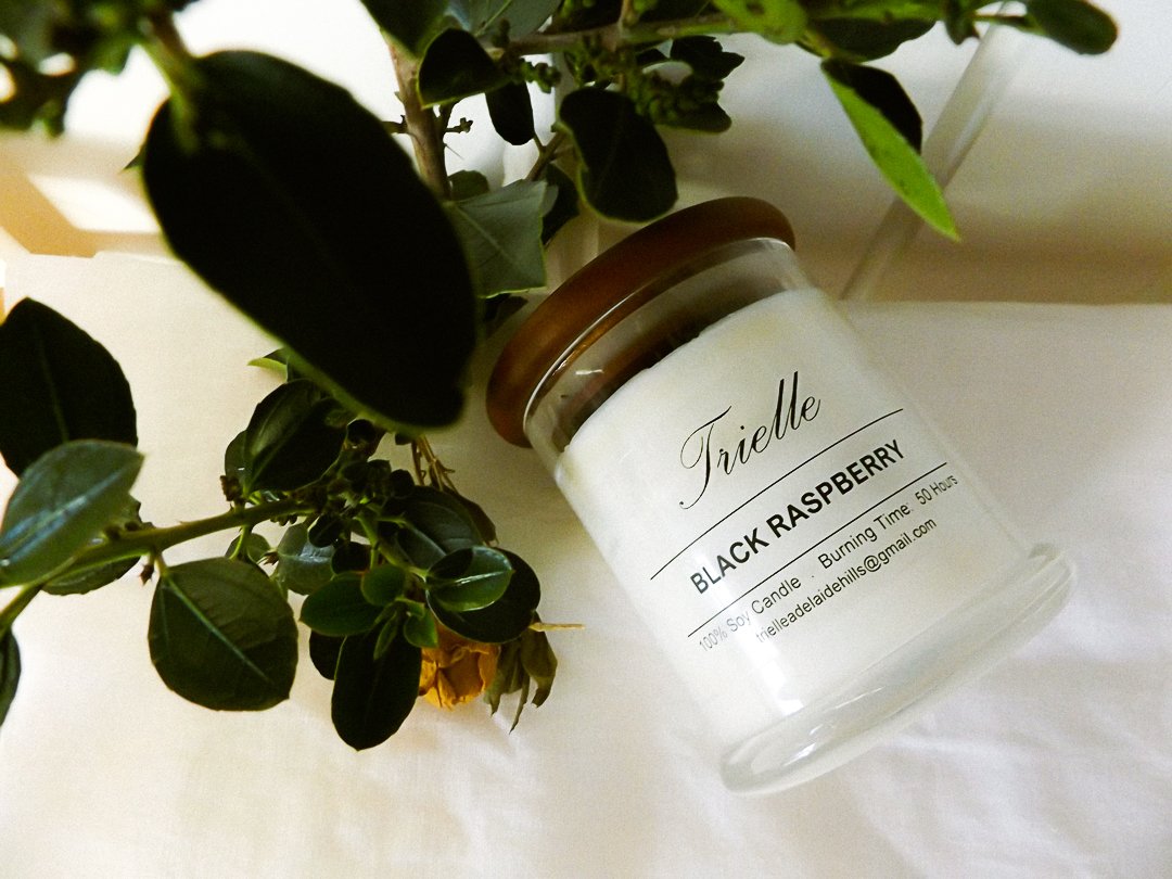 A white candle labeled 'Trielle Black Raspberry' next to a green plant with yellow flowers.