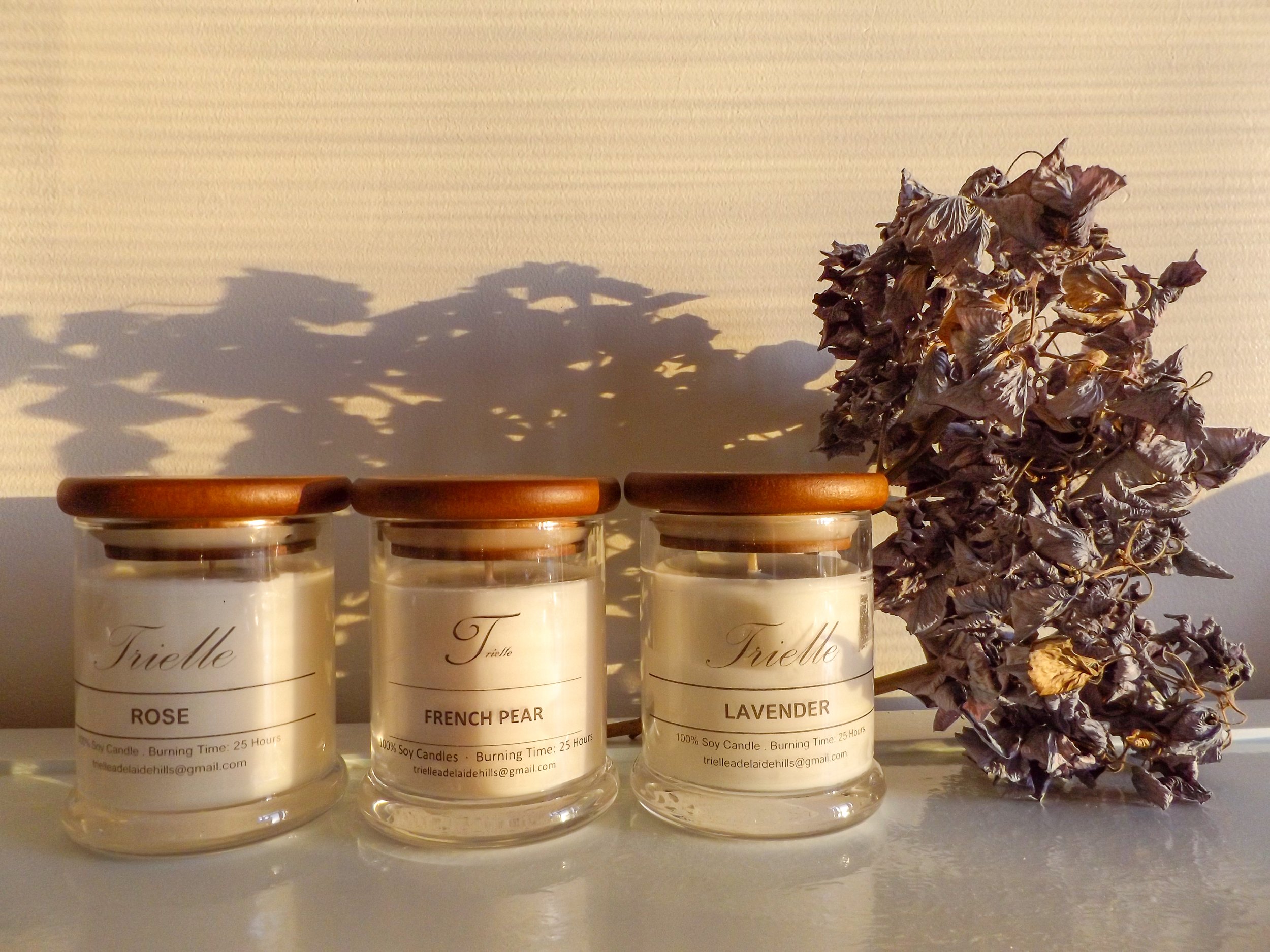 Three candles labeled rose, French pear, and lavender in glass jars positioned on a shelf with a dried lavender bouquet to the right. Shadows cast on a light-colored wall behind them.