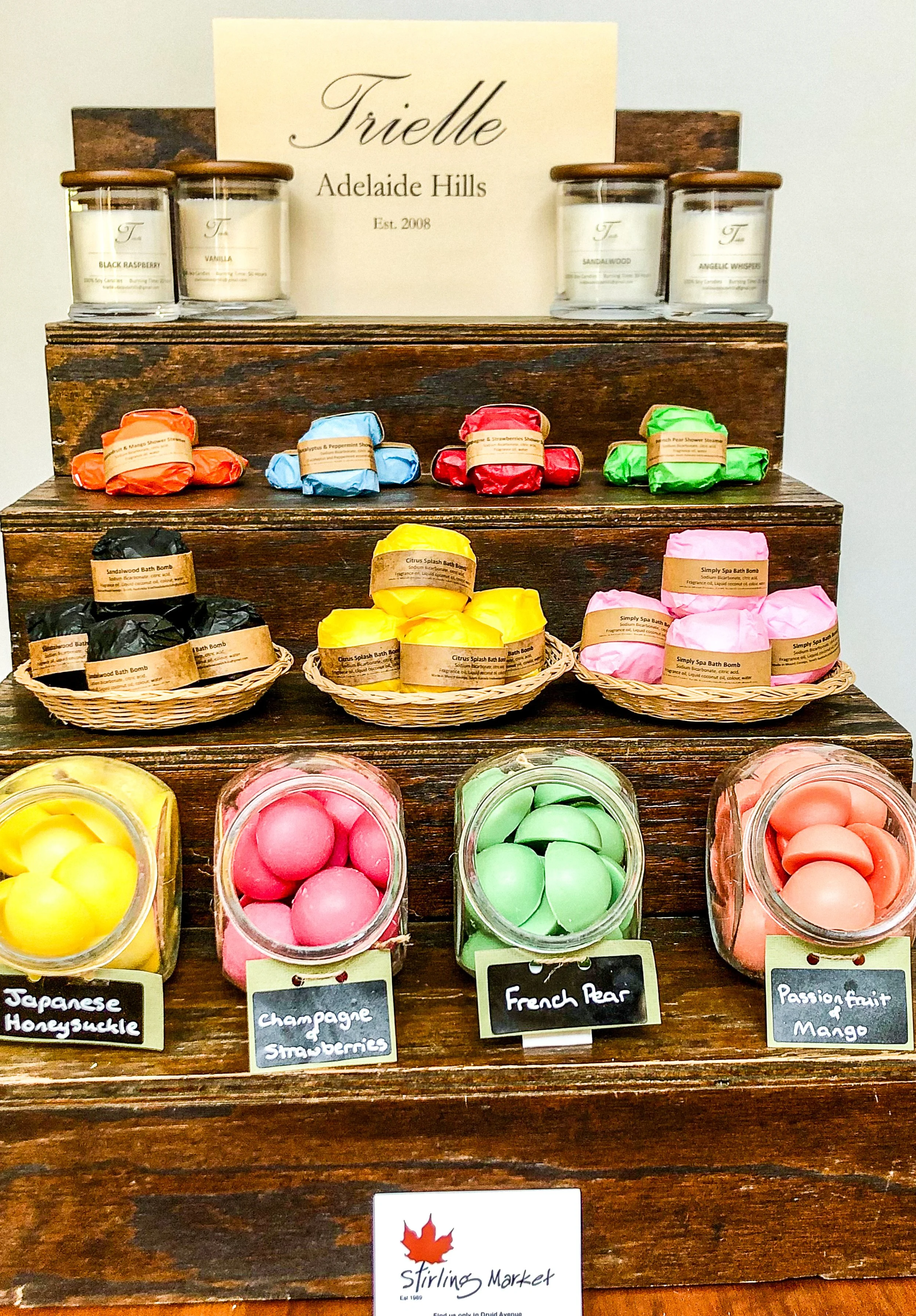 Display of colorful bath bombs and candles on a wooden shelf at Stirling Market. The candles in glass jars with wooden lids are labeled with scents: Black Raspberry, Vanilla, Sandalwood, and Angelic Wisteria. The bath bombs are arranged in baskets with labels: Japanese Honeysuckle, Champagne and Strawberries, French Pear, and Passion Fruit & Mango.
