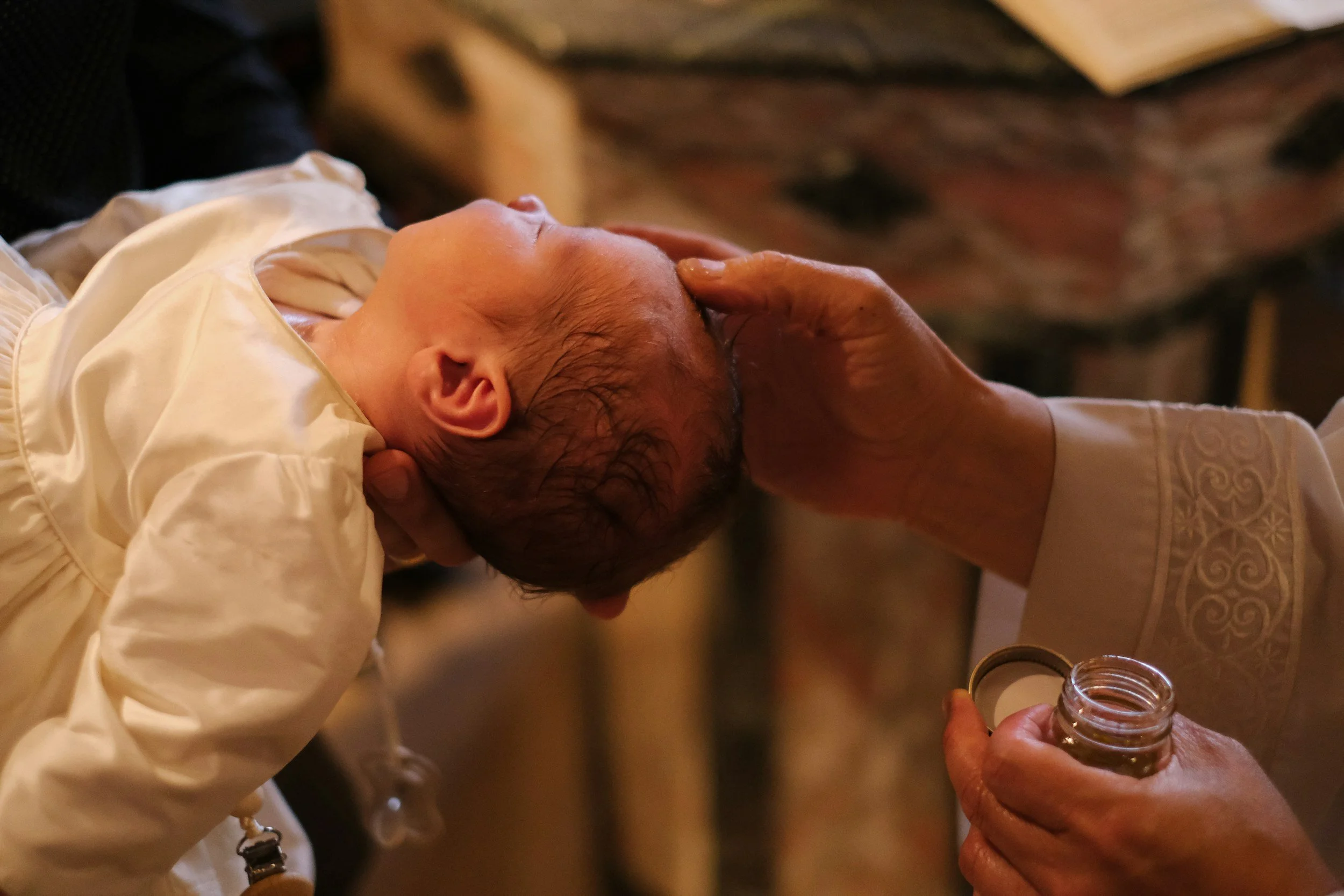 A person holding a baby close, with the baby's face resting on the person's chest, in a warm indoor setting. An adult hand is holding a small jar of honey, and another adult's hand is tenderly cradling the baby's head.