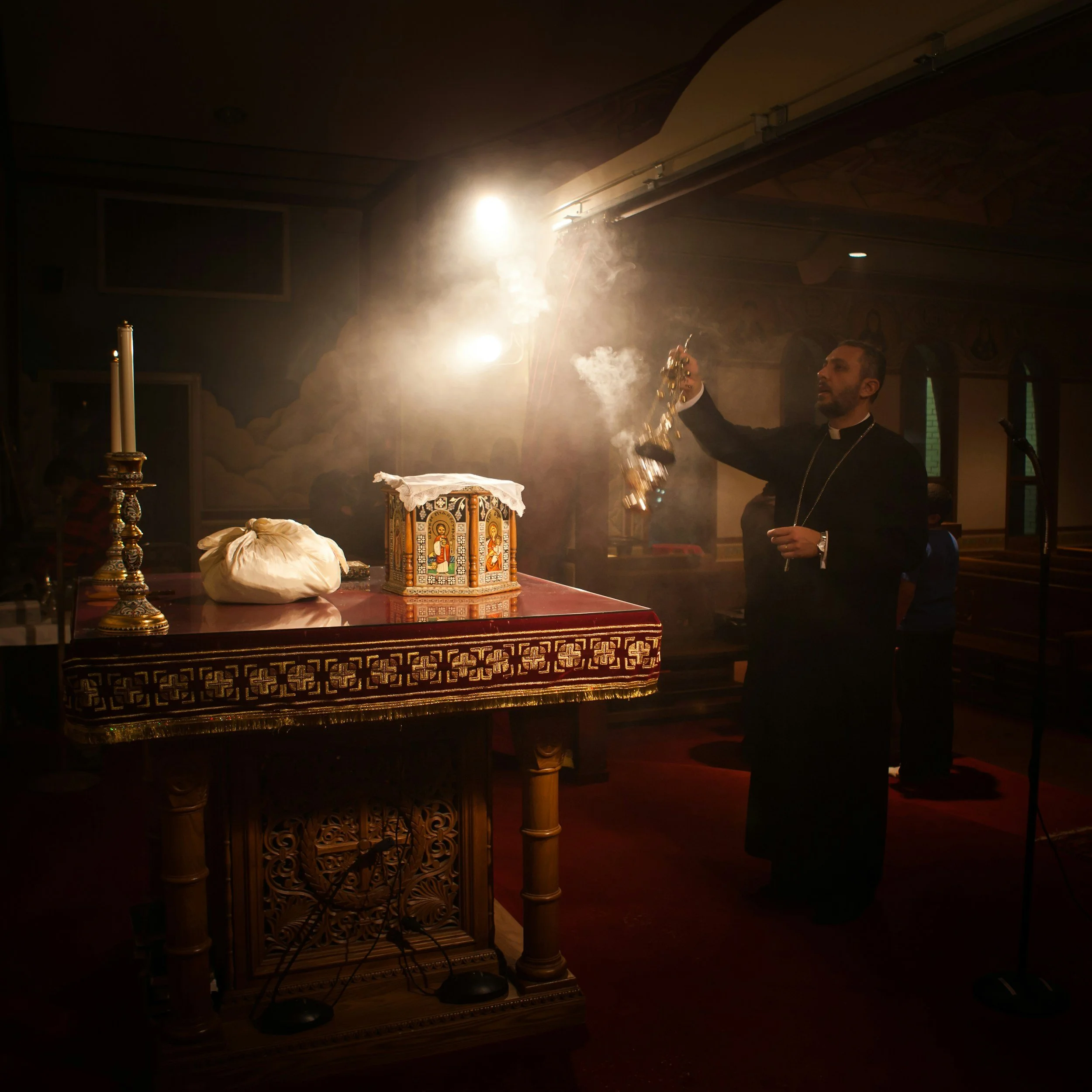 A priest holding a censer during a religious ceremony inside a church, with lit candles and religious icons in the background.