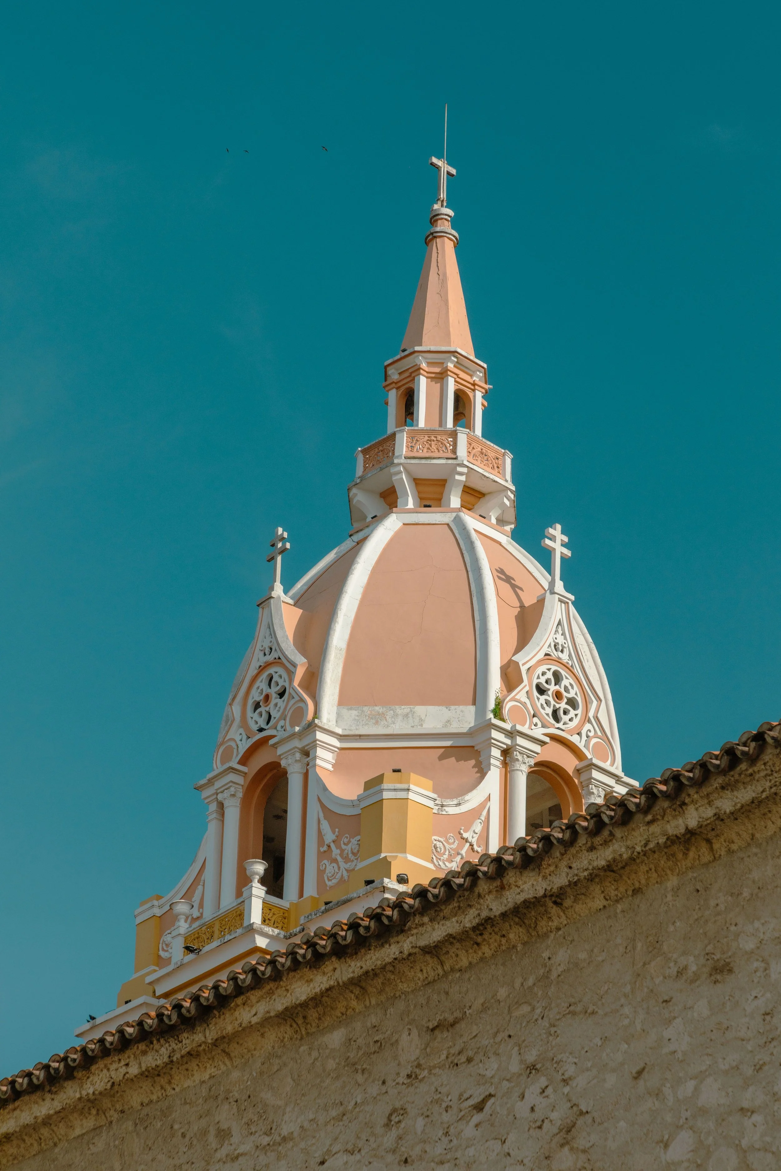 A peach-colored church tower with ornate white and gold architectural details, topped with a cross and a spire, against a blue sky with a few birds.
