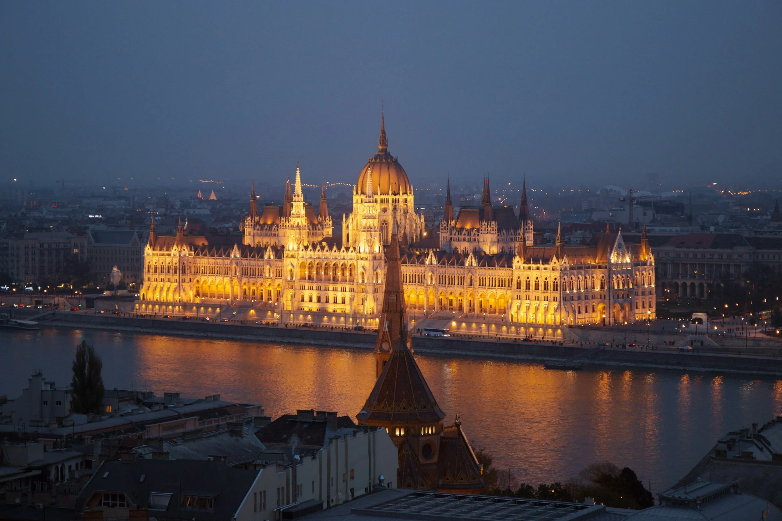 Nighttime view of the Hungarian Parliament Building illuminated along the riverbank in Budapest, Hungary, with city lights in the background.