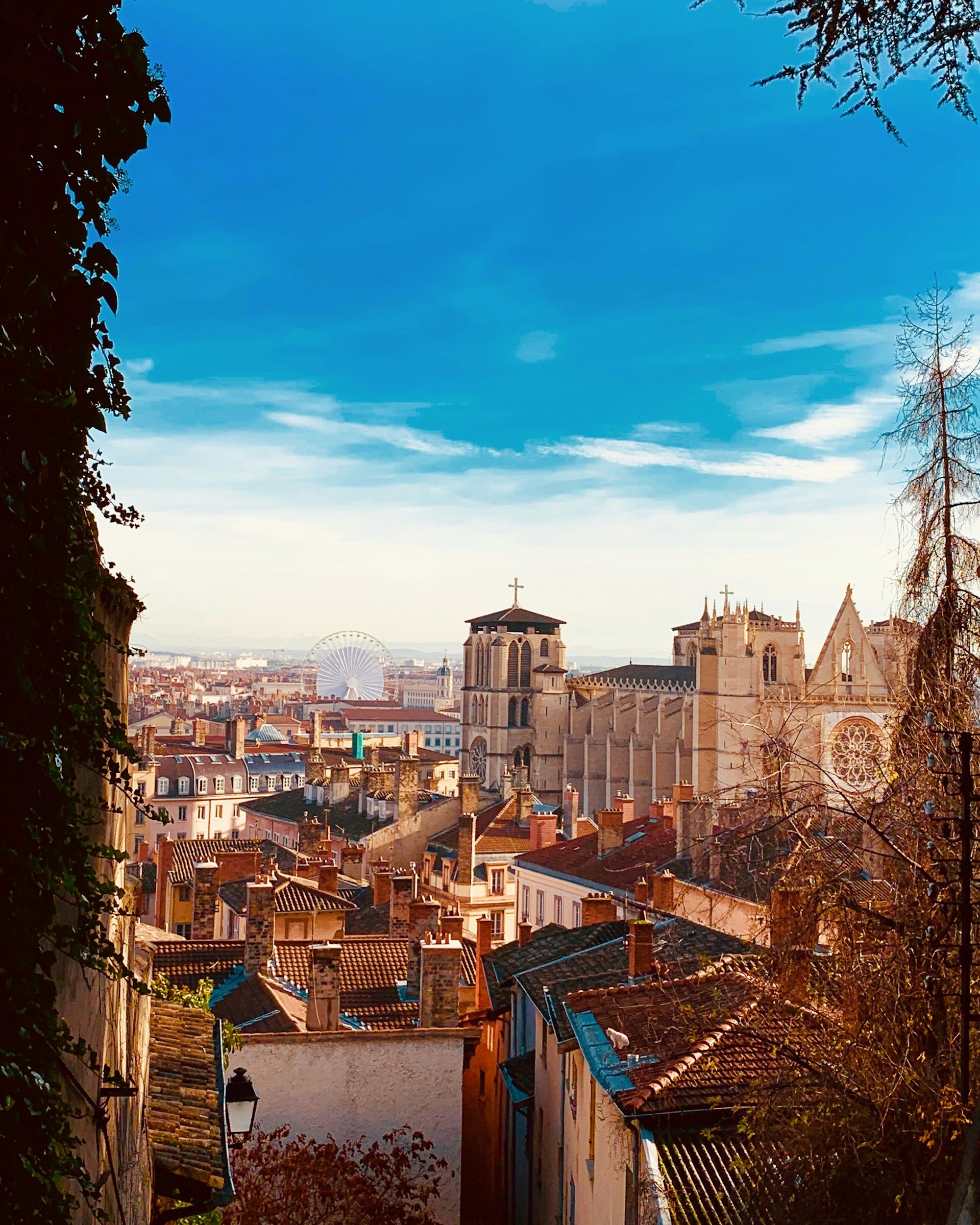 View of a city with old buildings, a church with a large stained glass window and a bell tower, a Ferris wheel in the distance, and a clear blue sky with a few clouds.