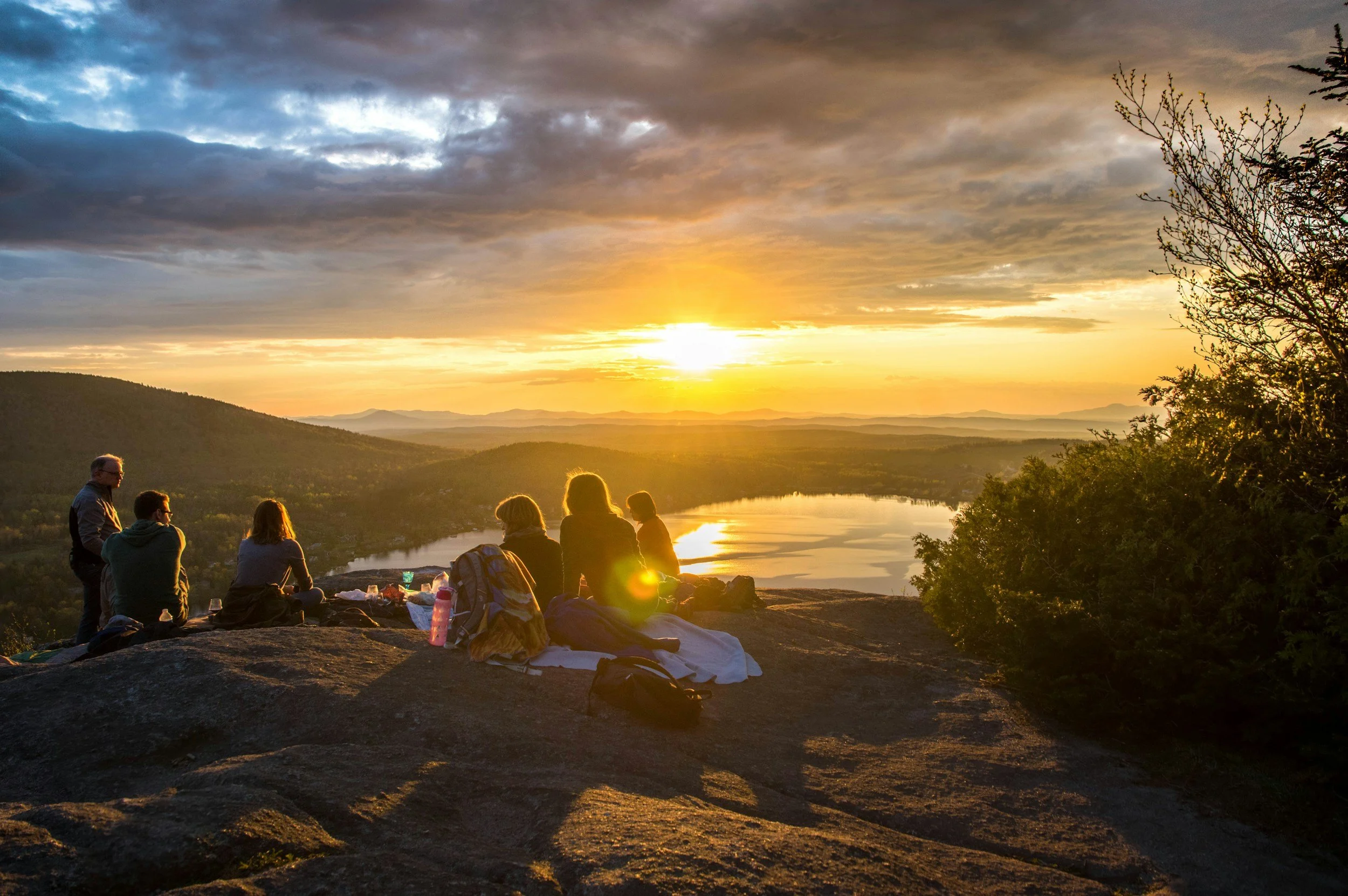 Group of people sitting outdoors on rocks watching sunset over a lake and mountains, some standing and some seated with backpacks and water bottles, surrounded by trees and a cloudy sky.