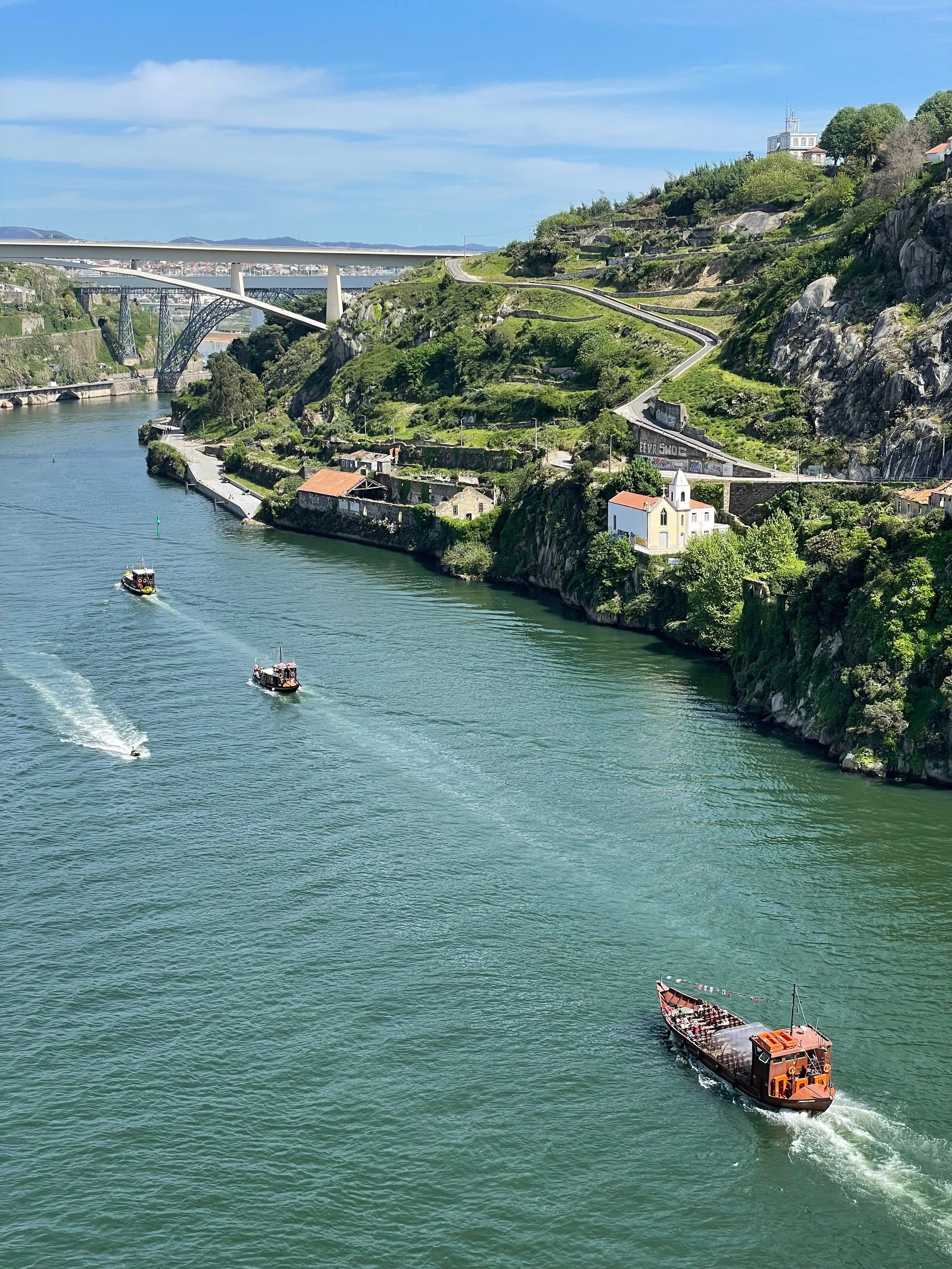 Ships sailing on a river with hillside buildings and bridges in the background.