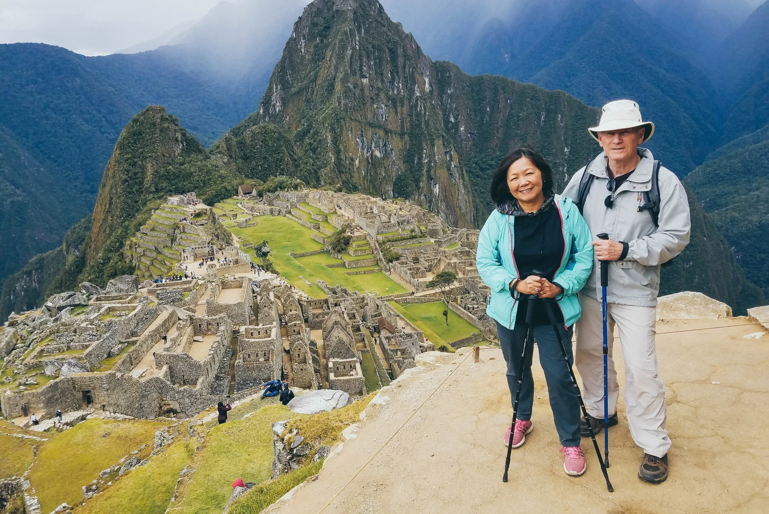 A woman and a man standing together in front of Machu Picchu, an ancient Incan city with stone ruins and terraces, surrounded by lush green mountains.