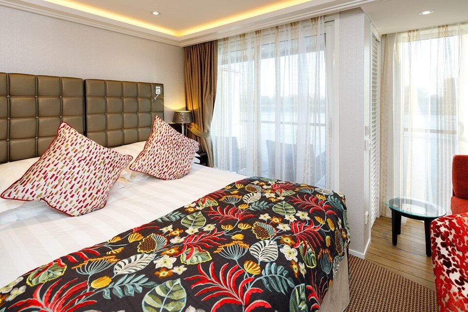 A hotel room with a large bed, colorful floral bedding, and four pillows, next to large windows with sheer curtains and a view outside.