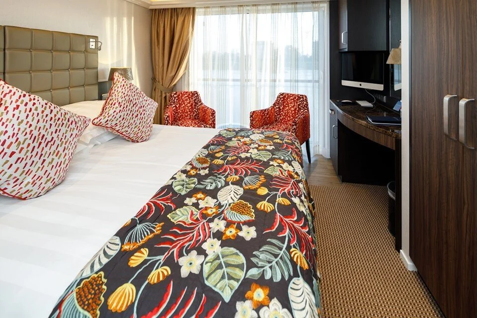 Hotel room with a bed, colorful bedspread, pillows, two red patterned chairs, a TV, and a window with curtains.