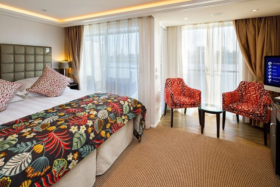 Hotel room with a bed, red patterned chairs, a small table, and a television, with large windows covered by sheer curtains and heavy drapes.