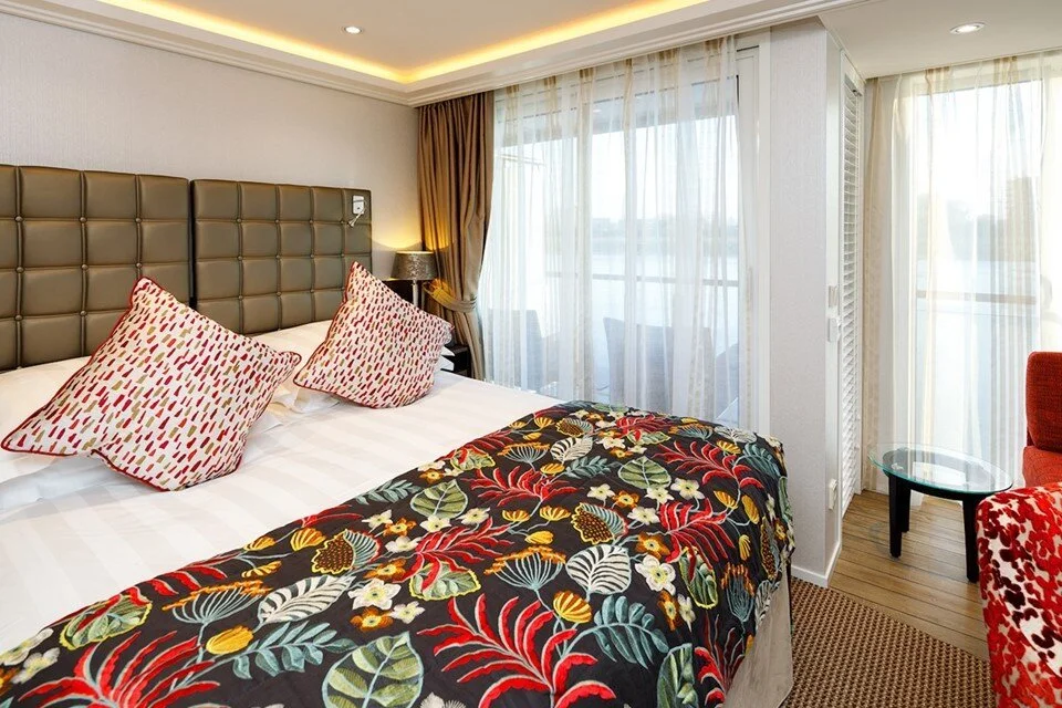 Hotel room with a bed, colorful floral blanket, two patterned pillows, nightstand with lamp, large window with sheer curtains, and a small round glass table near a red chair.
