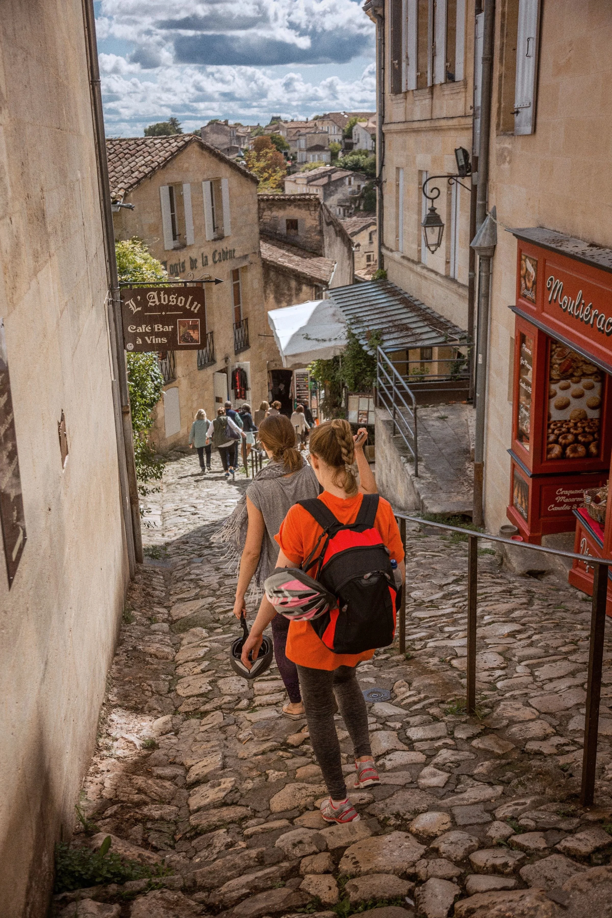 People walking down a narrow, cobblestone alley in a historic European town with stone buildings and shops, under a partly cloudy sky.