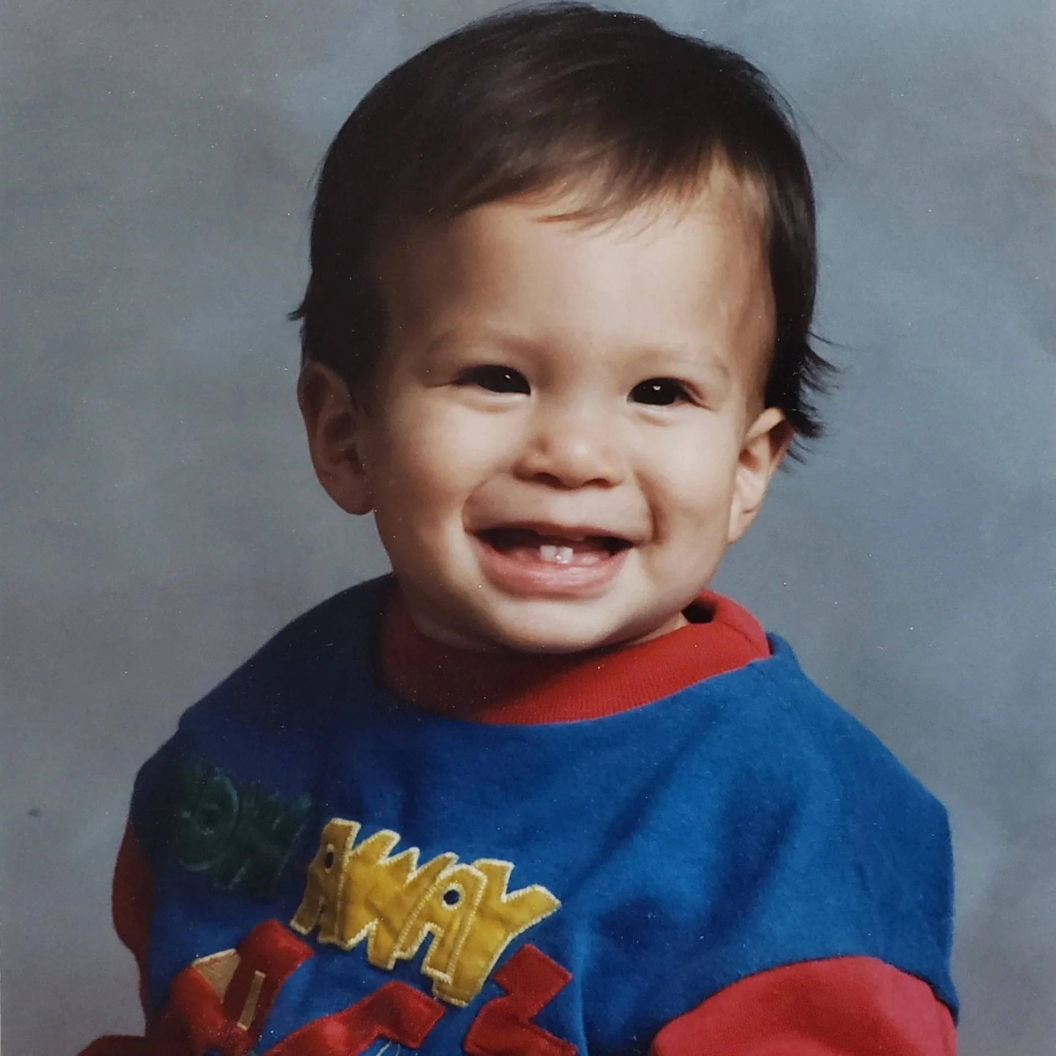Close-up of a young boy with dark hair, smiling, wearing a blue and red cap and a blue shirt with yellow design.