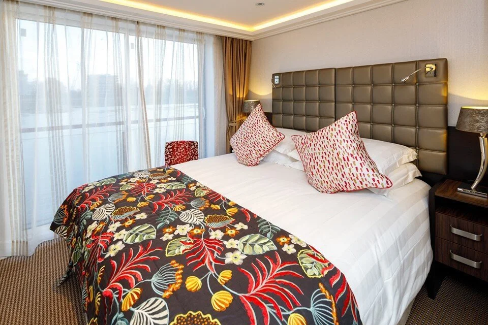 Hotel room with a bed, decorative pillows, a patterned bed runner, bedside lamps, a window with sheer curtains, and a small chair.