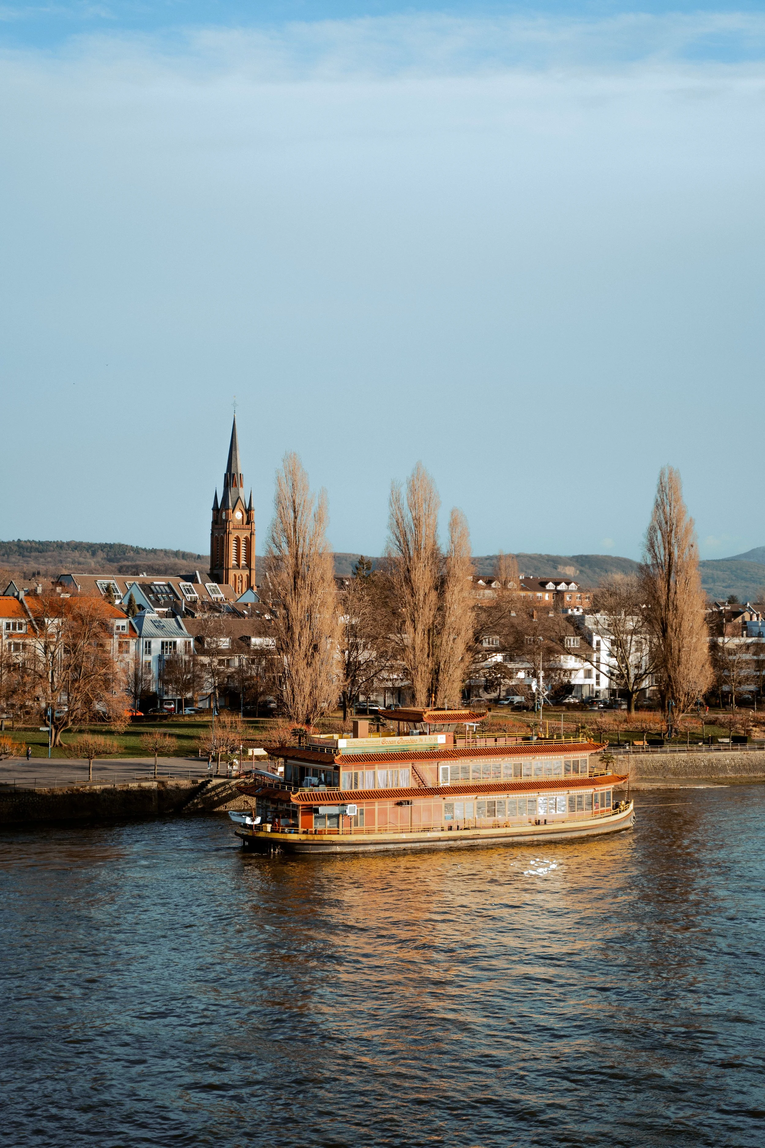 A river with a floating restaurant, a town with houses, a church with a tall steeple, trees, and hills in the background under a partly cloudy sky.