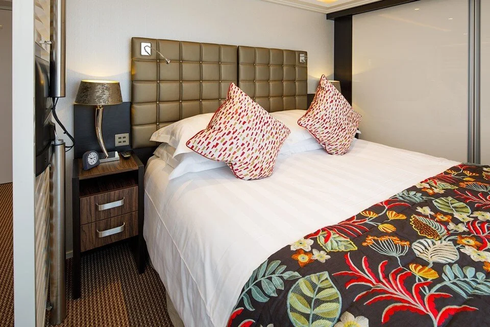 A neatly made hotel bed with a padded headboard, two patterned pillows, and a floral bed runner. There is a nightstand with a lamp, clock, and a wall-mounted reading light beside the bed.