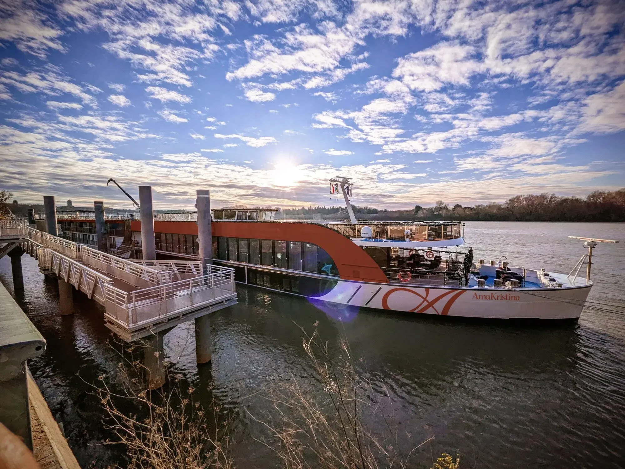 A boat named Anna Kristina docked along a river with a walkway, with a partly cloudy sky and the sun setting in the background.