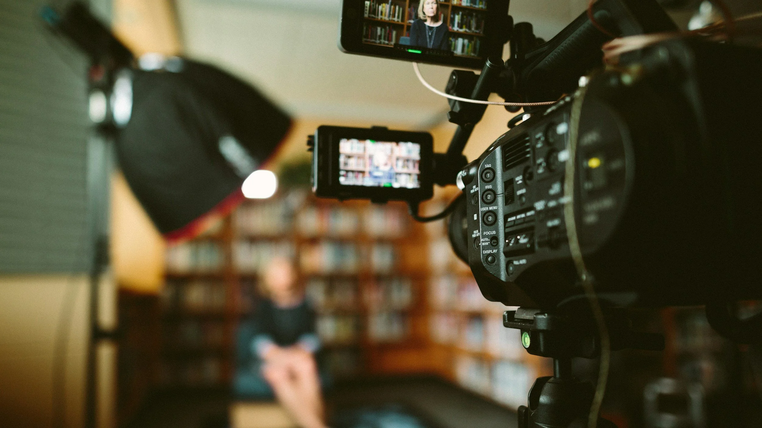Camerawork capturing a woman in a library being interviewed or recorded, with one camera focused on her and a second camera capturing the scene, both pointing towards the woman with bookshelves in the background.