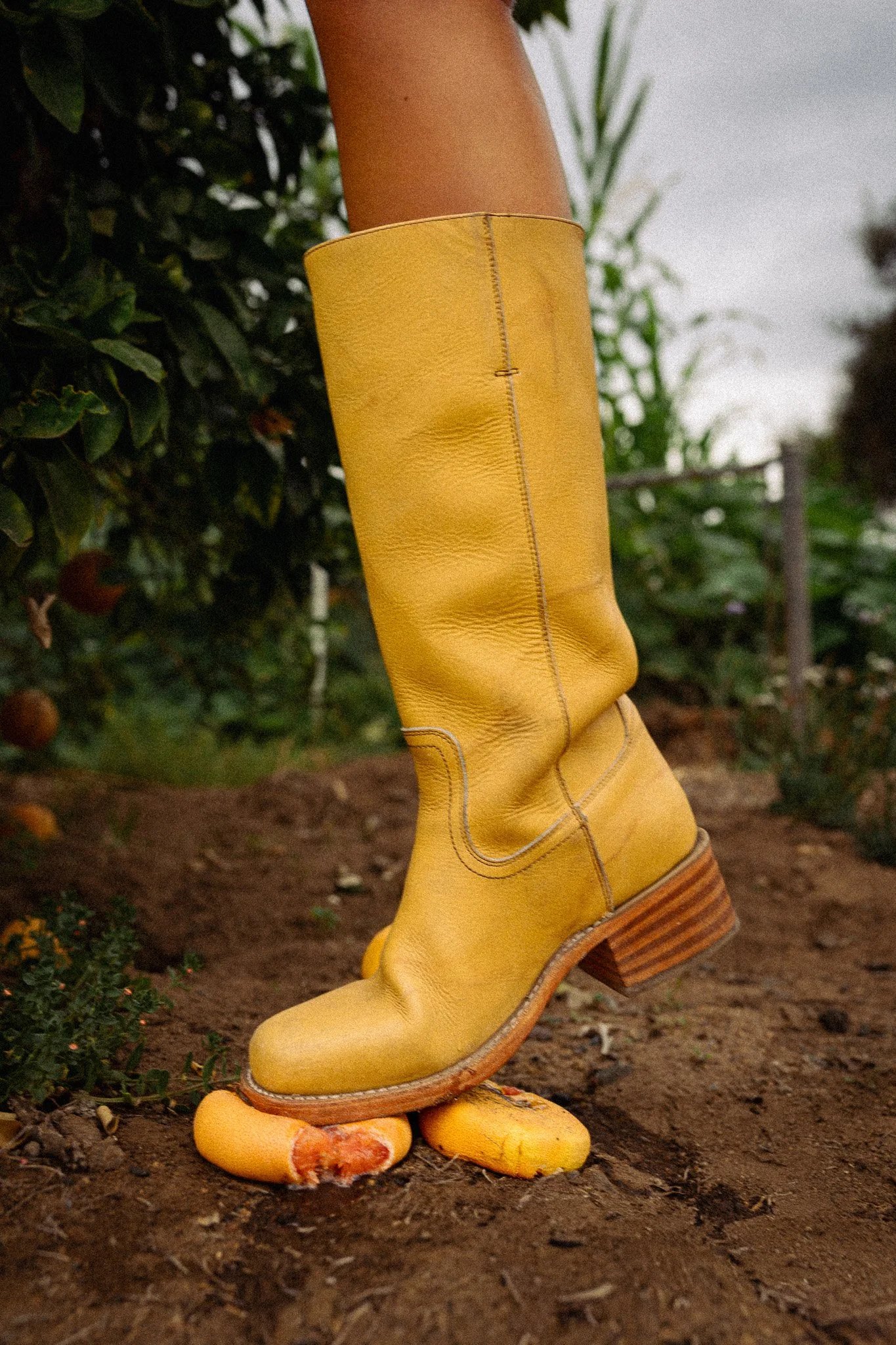 A person wearing yellow boots walking on dirt ground outdoors, with green plants and a cloudy sky in the background.