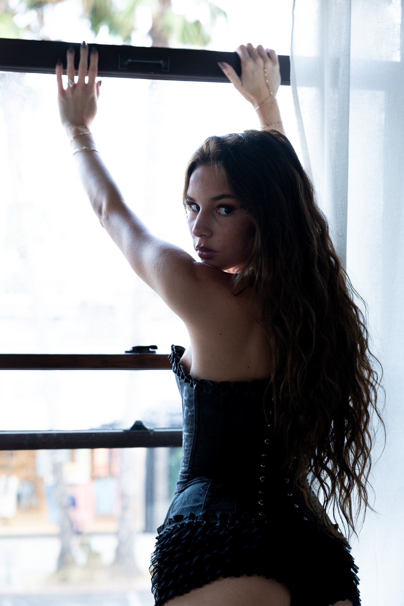A woman with long wavy hair standing by a window, holding the window frame with her hands, looking at the camera with a serious expression, wearing a black strapless dress with ruffles.