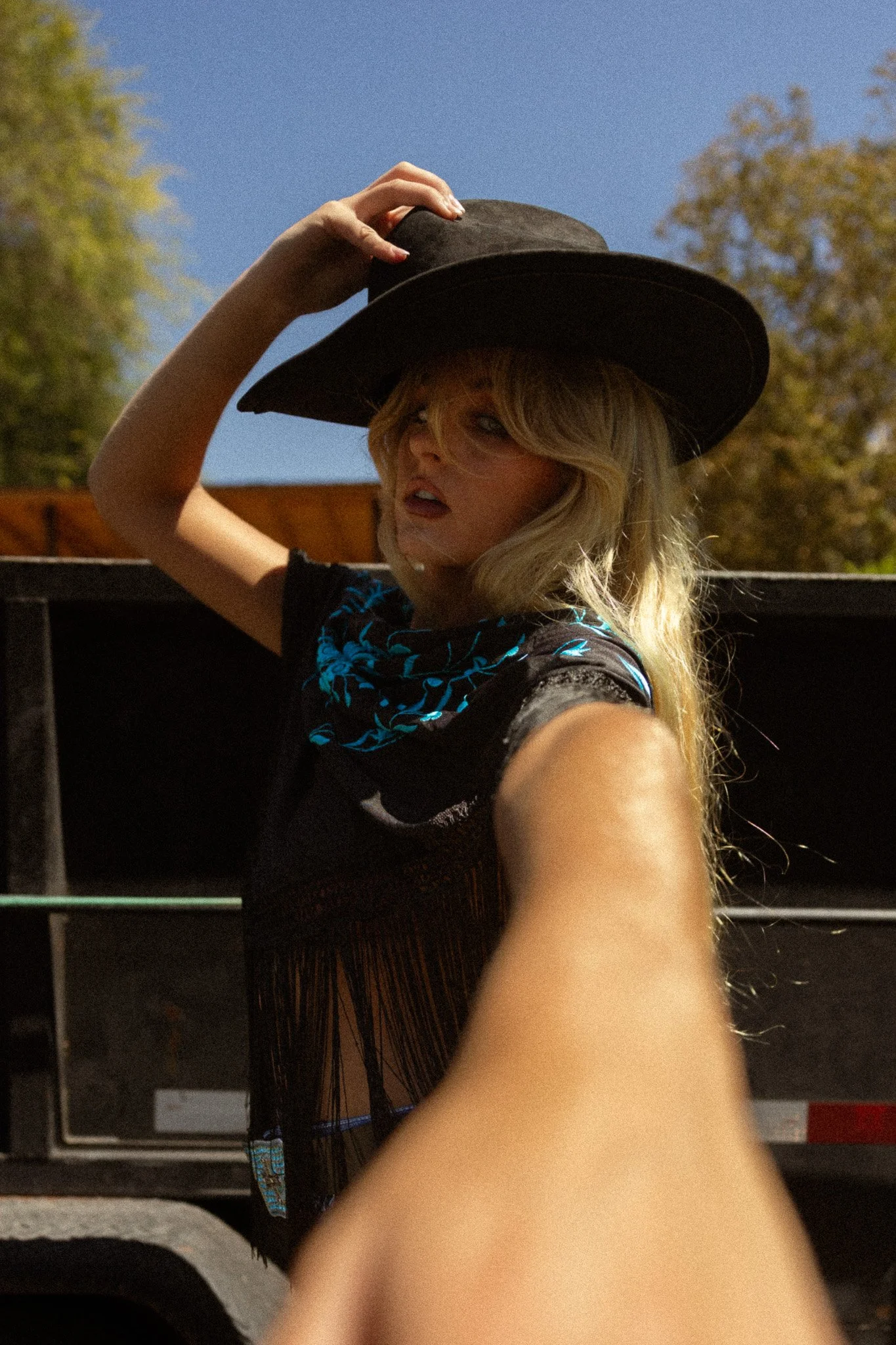 A woman with blonde hair and glasses wearing a black hat, a black shirt with blue designs, taking a selfie outdoors during daytime, with trees and a blue sky in the background.