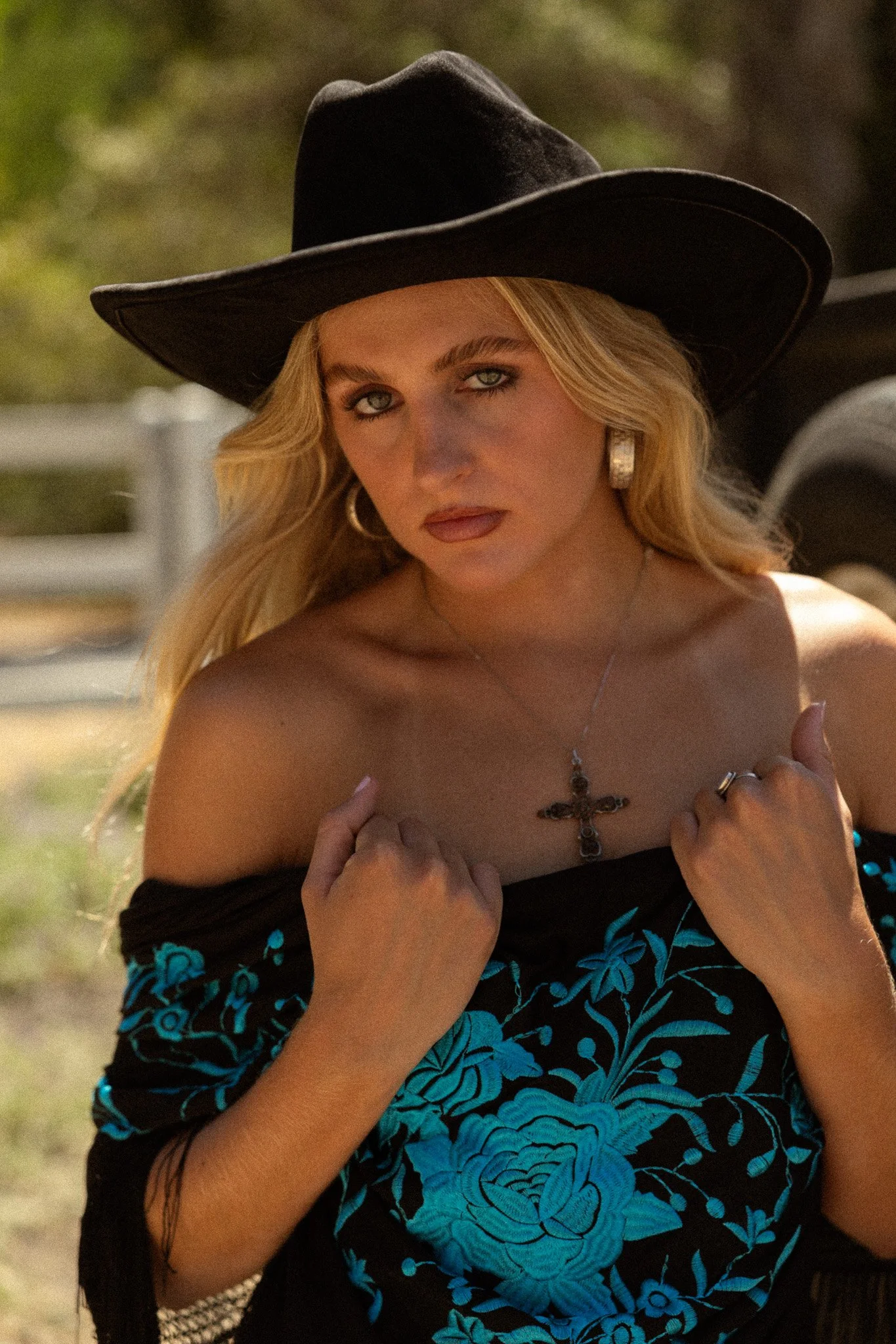 A woman with long blonde hair, blue eyes, wearing large earrings and a black wide-brimmed hat. She is holding her off-the-shoulder black dress with blue floral embroidery, and wearing a necklace with a cross pendant.