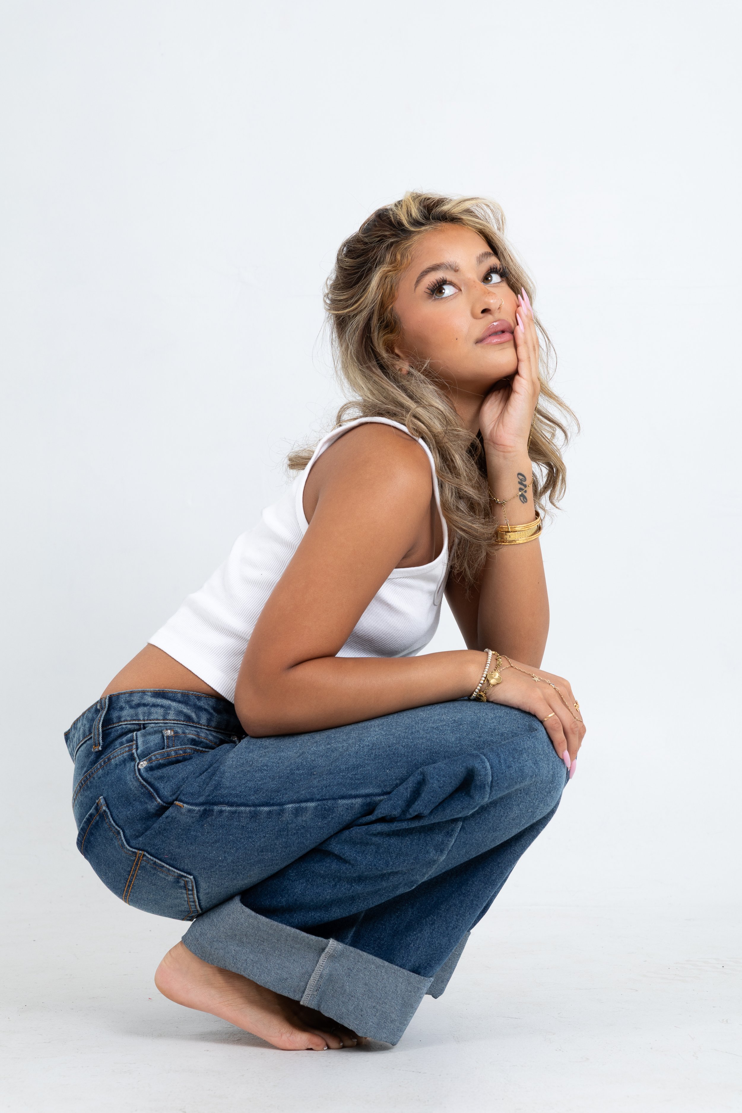 A woman with long wavy blonde hair, wearing a white tank top and blue jeans, is squatting with her right arm resting on her knee, and her left hand touching her face, on a plain white background.