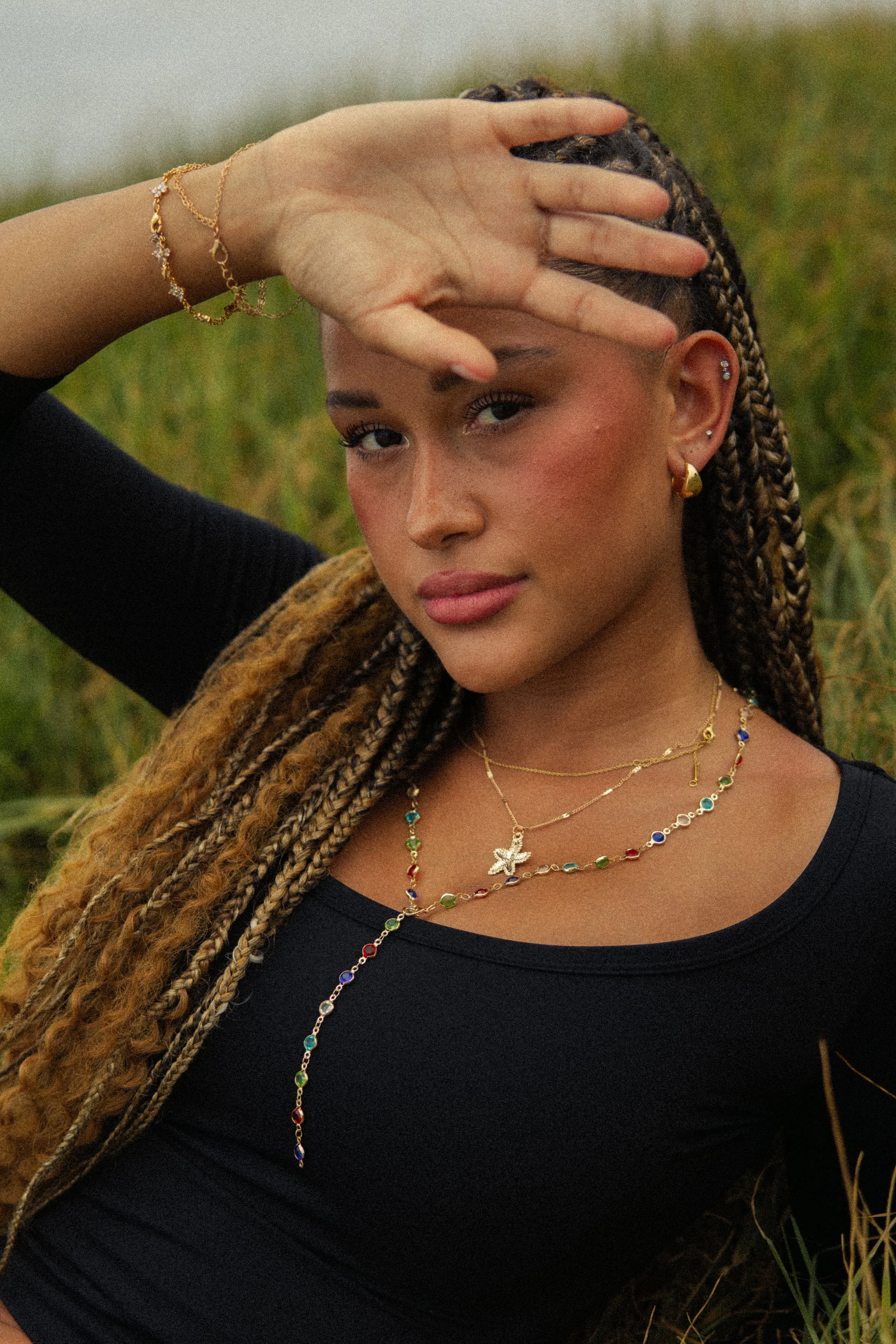 A woman with braided hair wearing gold jewelry and a black top, outdoors with grass in the background.