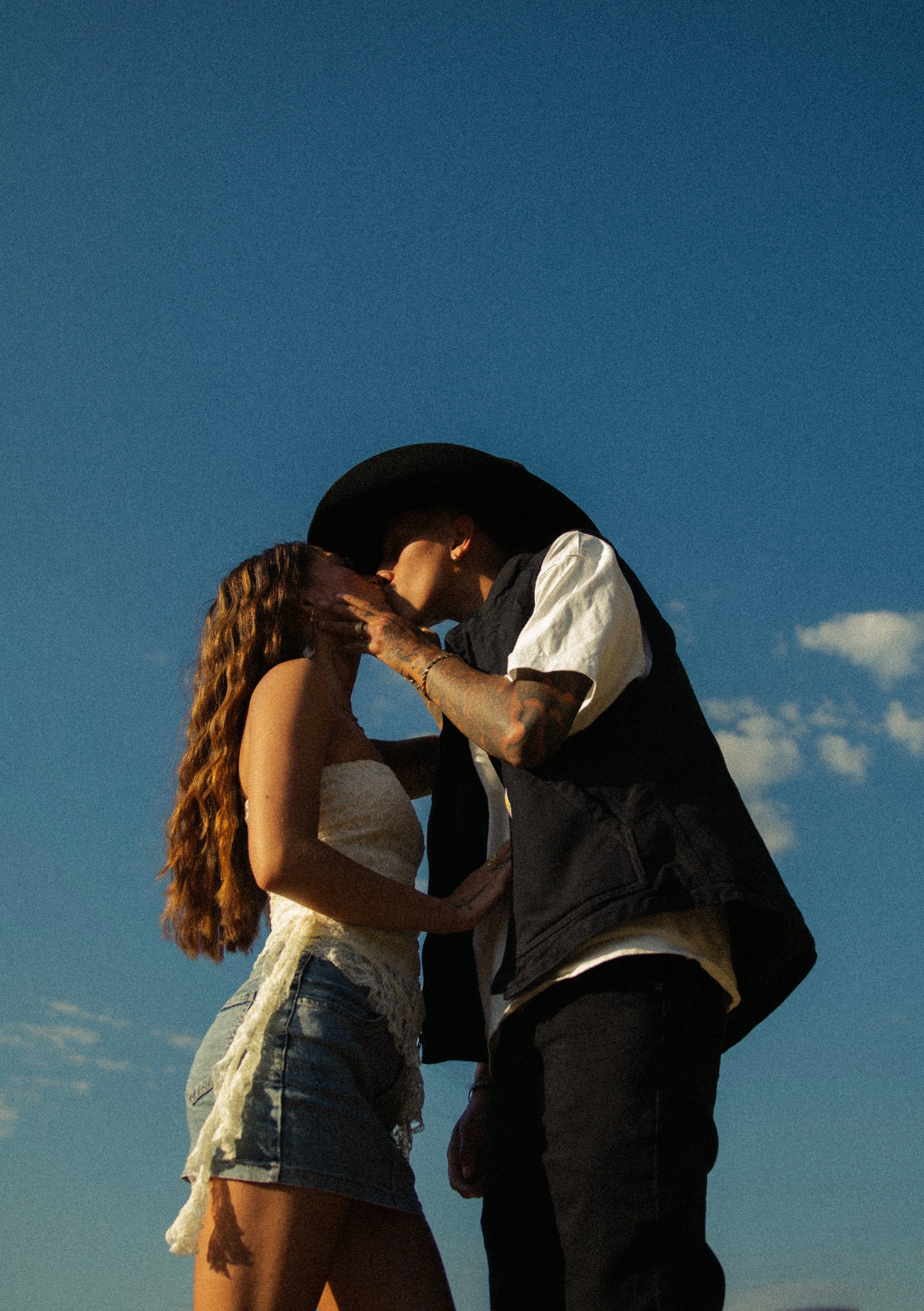 A couple stands close together, sharing a kiss against a clear blue sky at sunset. The woman has long, wavy hair and wears a strapless top and denim shorts. The man wears a black hat, black vest over a light shirt, and dark pants.