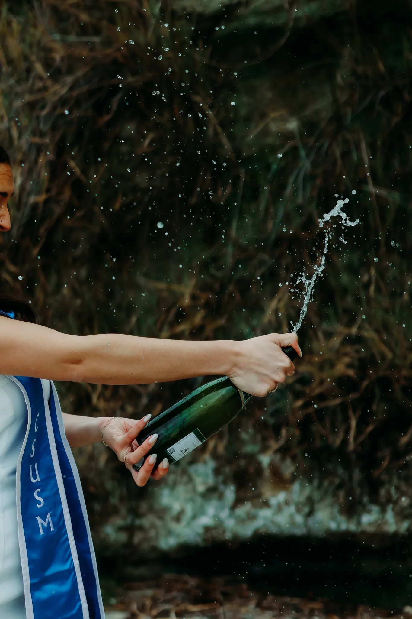 Person holding a champagne bottle, spraying it outdoors with a natural background, possibly near a waterfall or stream.