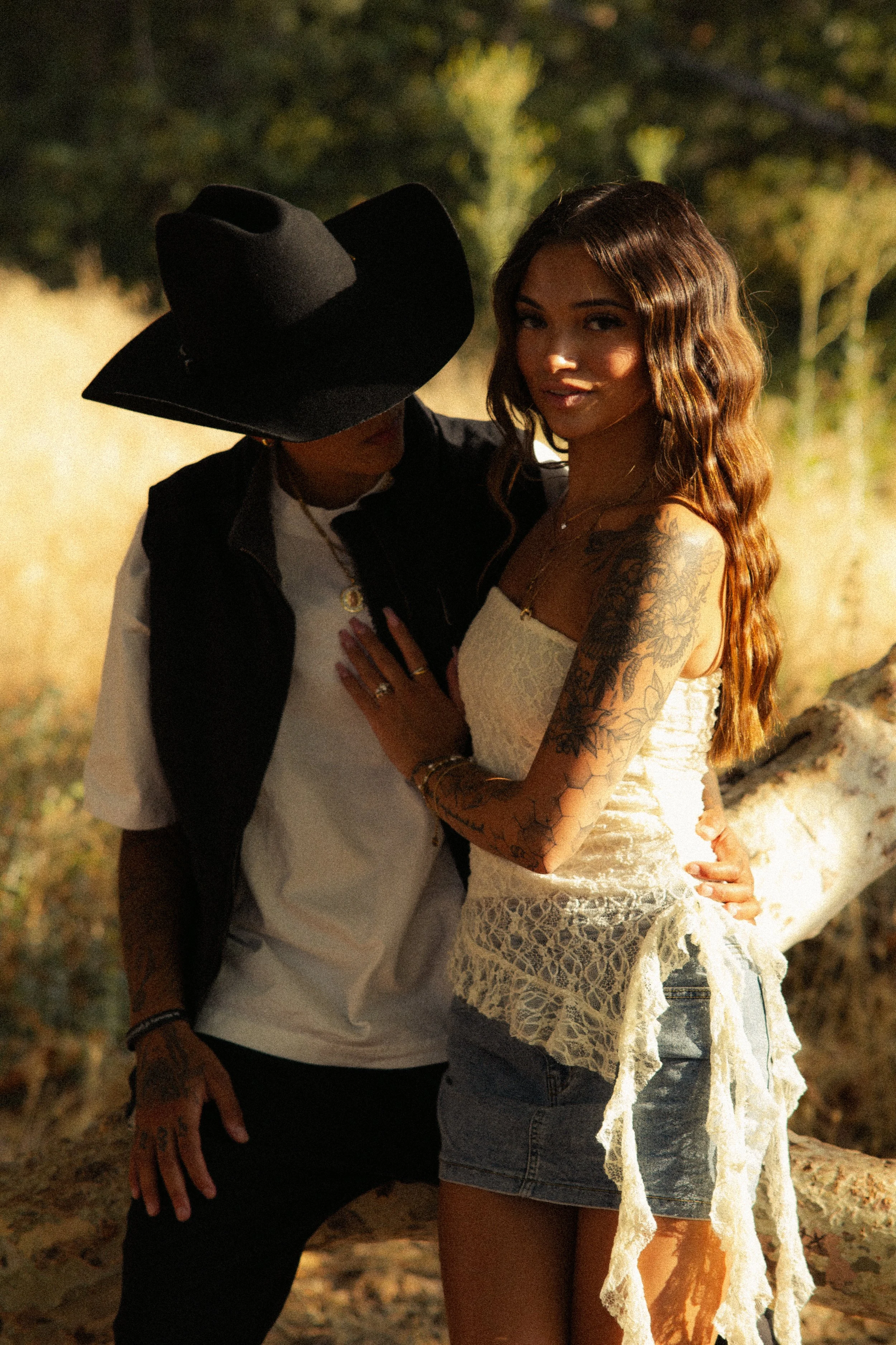 A woman in a white lace dress and denim shorts with long wavy hair standing next to a person wearing a large black hat and black vest, outdoors on a sunny day.