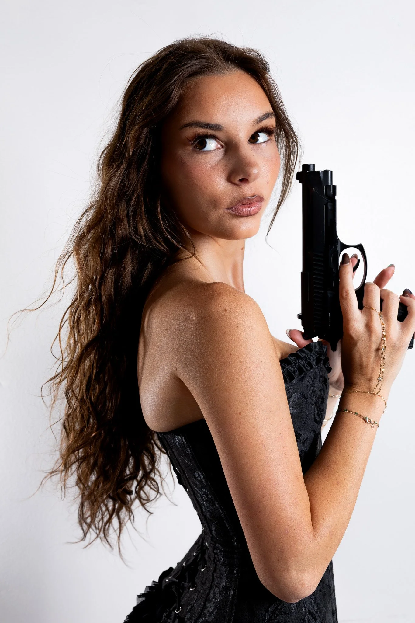 A woman with long wavy brown hair holding a black handgun, wearing a black lace dress, against a plain white background.