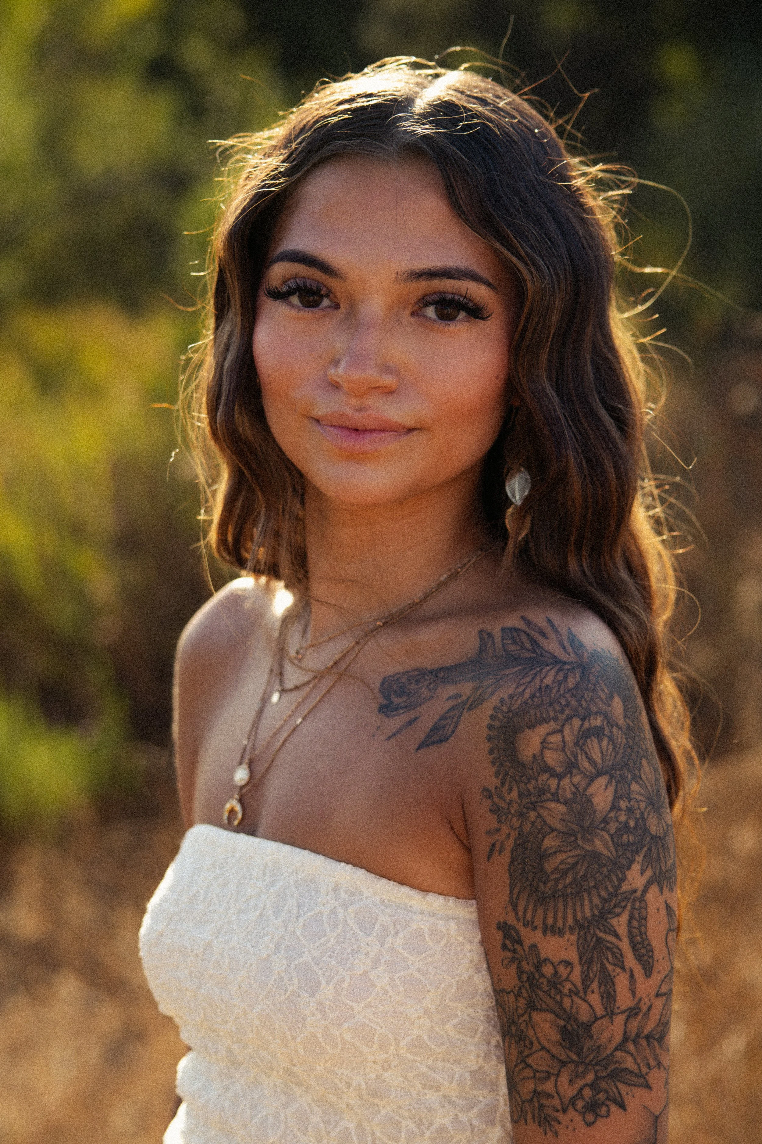 A woman with long wavy hair, dark eyeliner, and makeup, wearing jewelry, a tattoo on her shoulder, and a white strapless dress, outdoors during sunset with blurred greenery in the background.