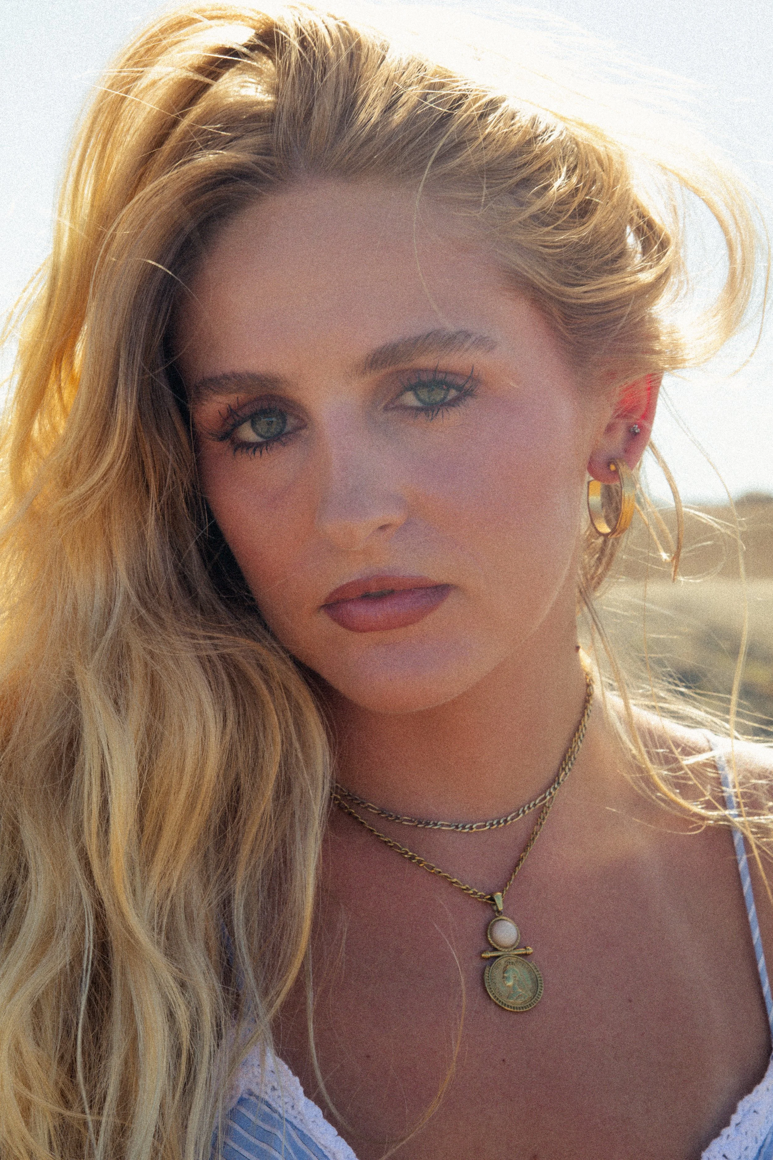 Close-up of a woman with long blonde hair, wearing gold jewelry, including layered necklaces and earrings, and a white top with a scalloped edge. The background appears to be outdoors with a bright sky.