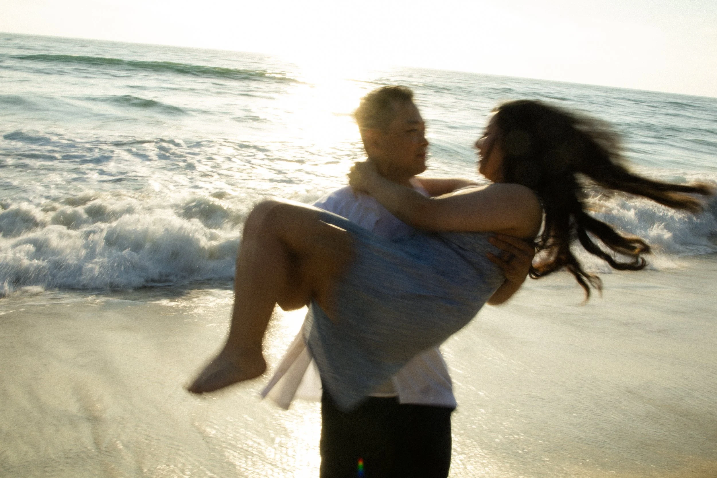 A couple on the beach, with the man holding the woman in his arms as they enjoy the sunset and waves.