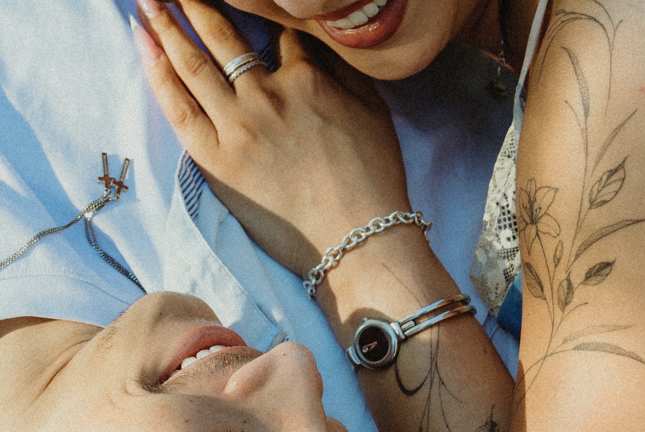 Close-up of a young woman smiling, lying on her side, with jewelry including rings, bracelets, and a watch, and tattoos of leaves and flowers on her arm.