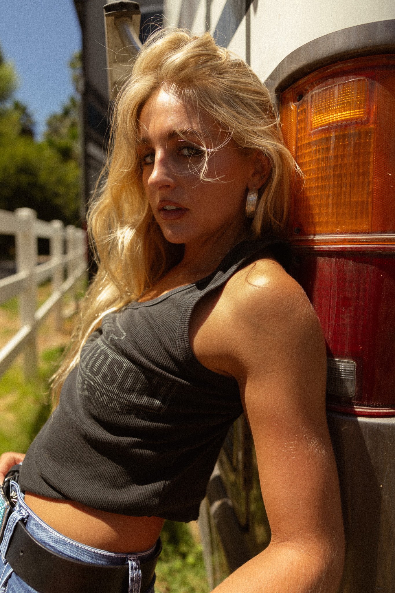 A blonde woman with wavy hair leaning against the rear of a red truck, wearing a dark tank top and jeans, outdoors with a white fence and green trees in the background.