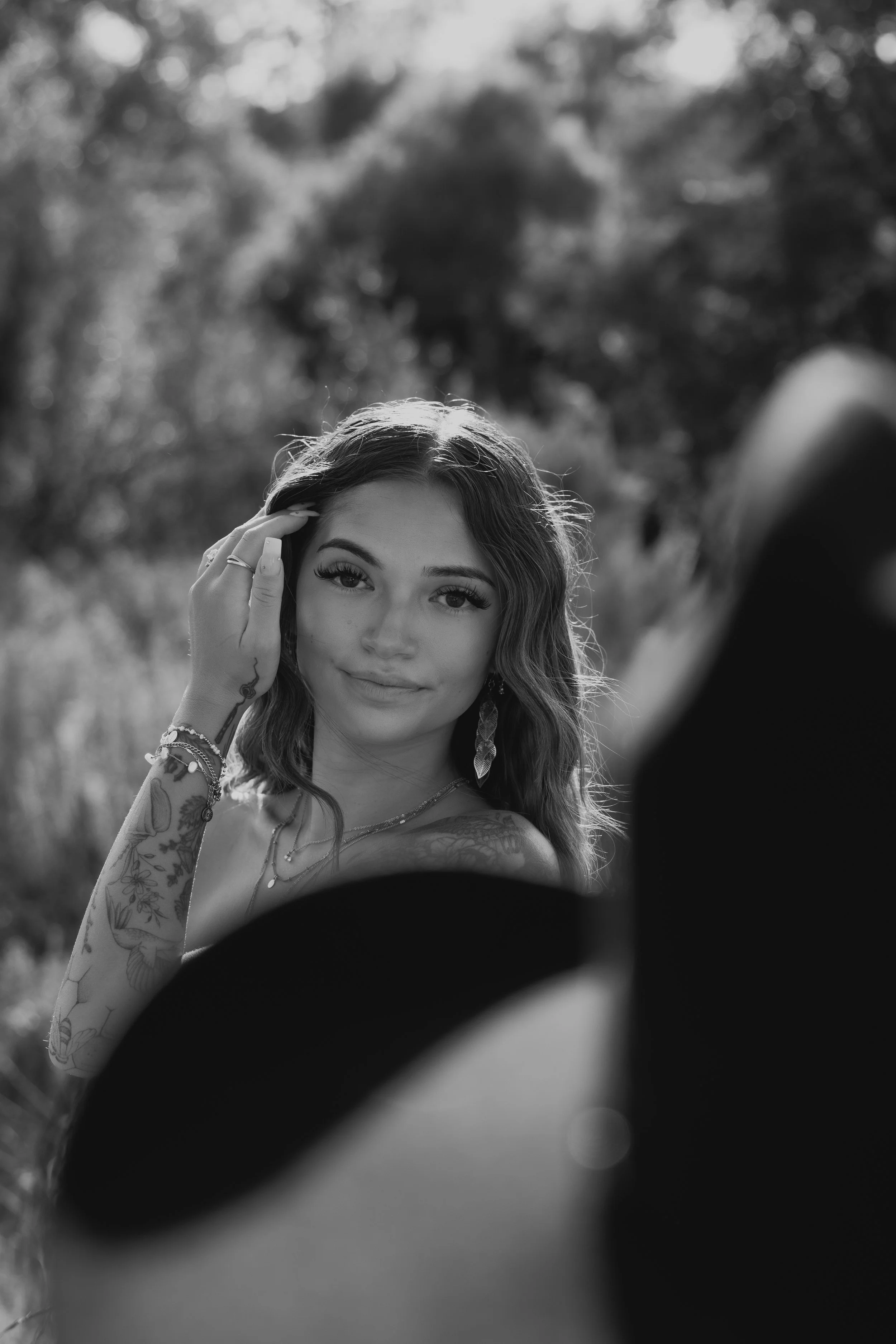 Black and white photo of a woman with long wavy hair, looking directly at the camera. She has a gentle smile, wears makeup, and is adorned with jewelry. The background is blurred with trees, suggesting an outdoor setting. The prompt appears to captur