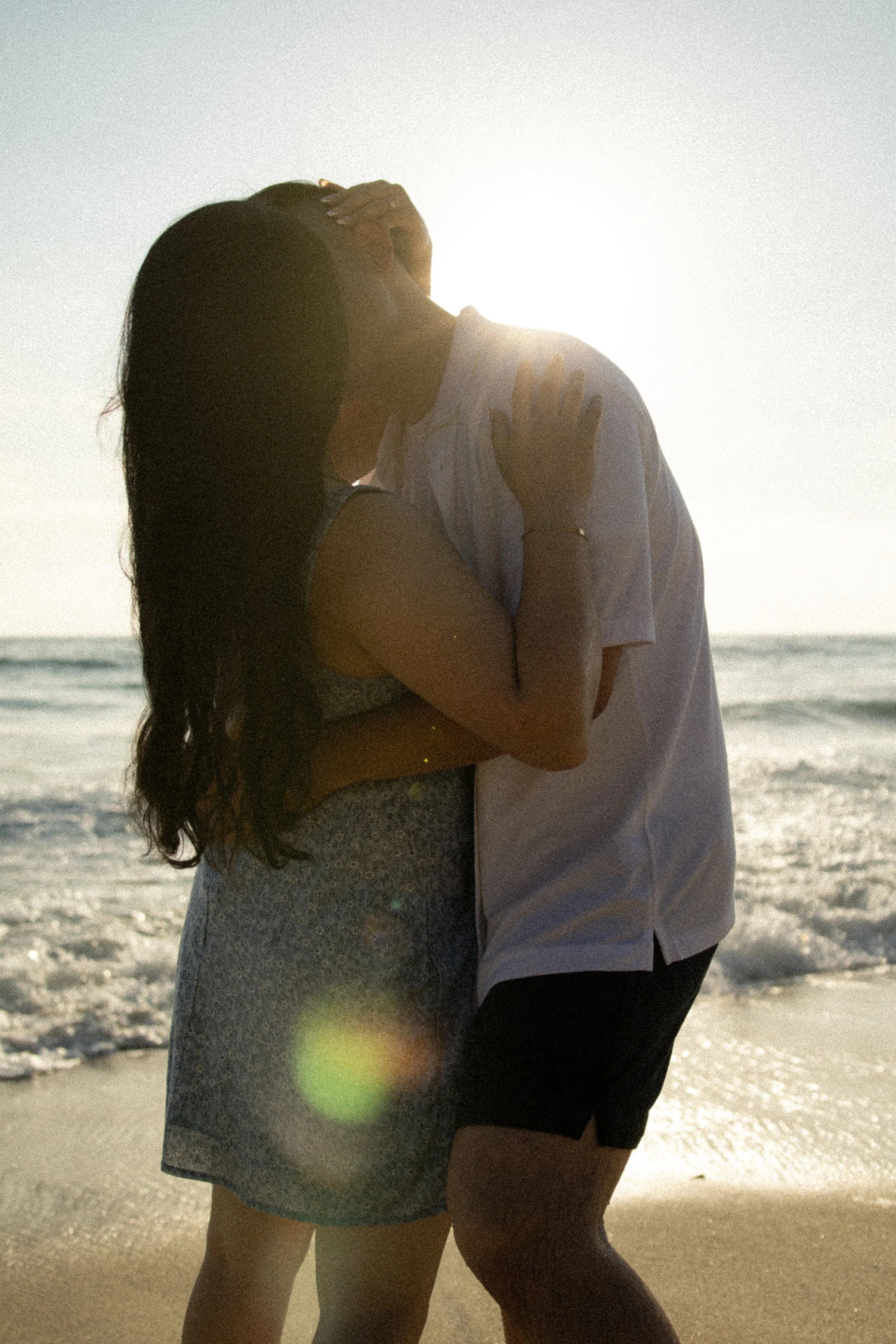 A couple hugging on the beach during sunset, their faces close together, with the ocean and sky in the background.
