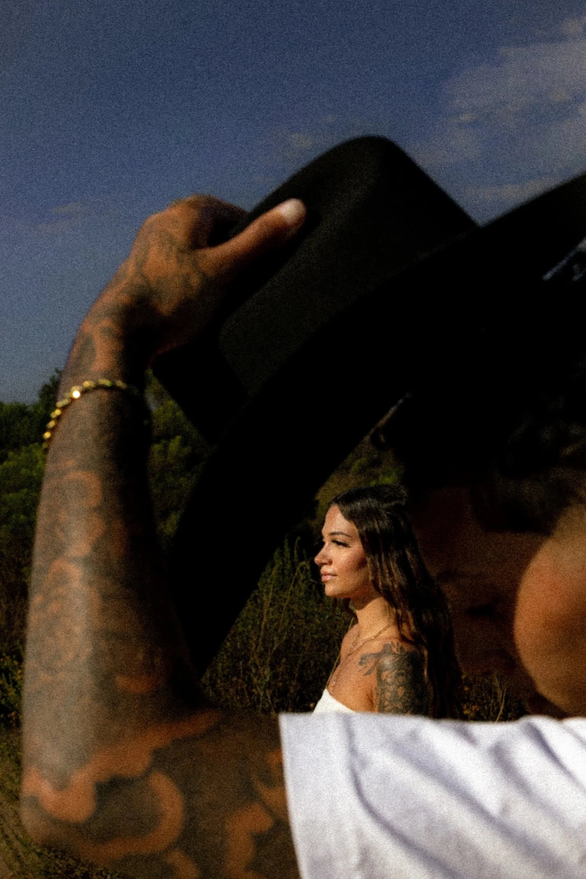 A woman with long brown hair and tattoos on her shoulder, standing outdoors during dusk or evening, appears to be posing for a photo. There is a person holding a black phone or camera in front of her.