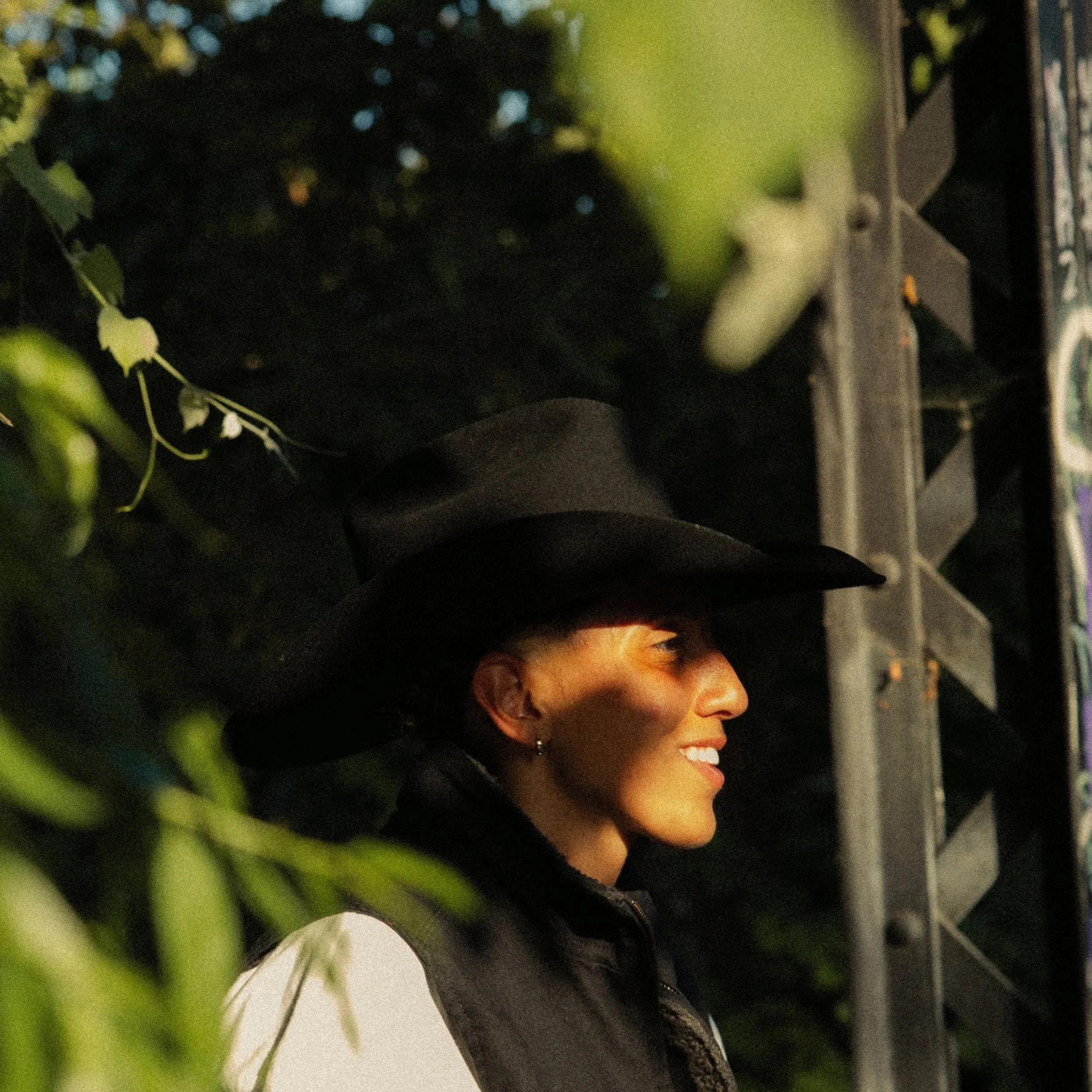 Side profile of a woman with curly black hair, wearing earrings and a black jacket, smiling and looking to her right at night with city lights in the background.