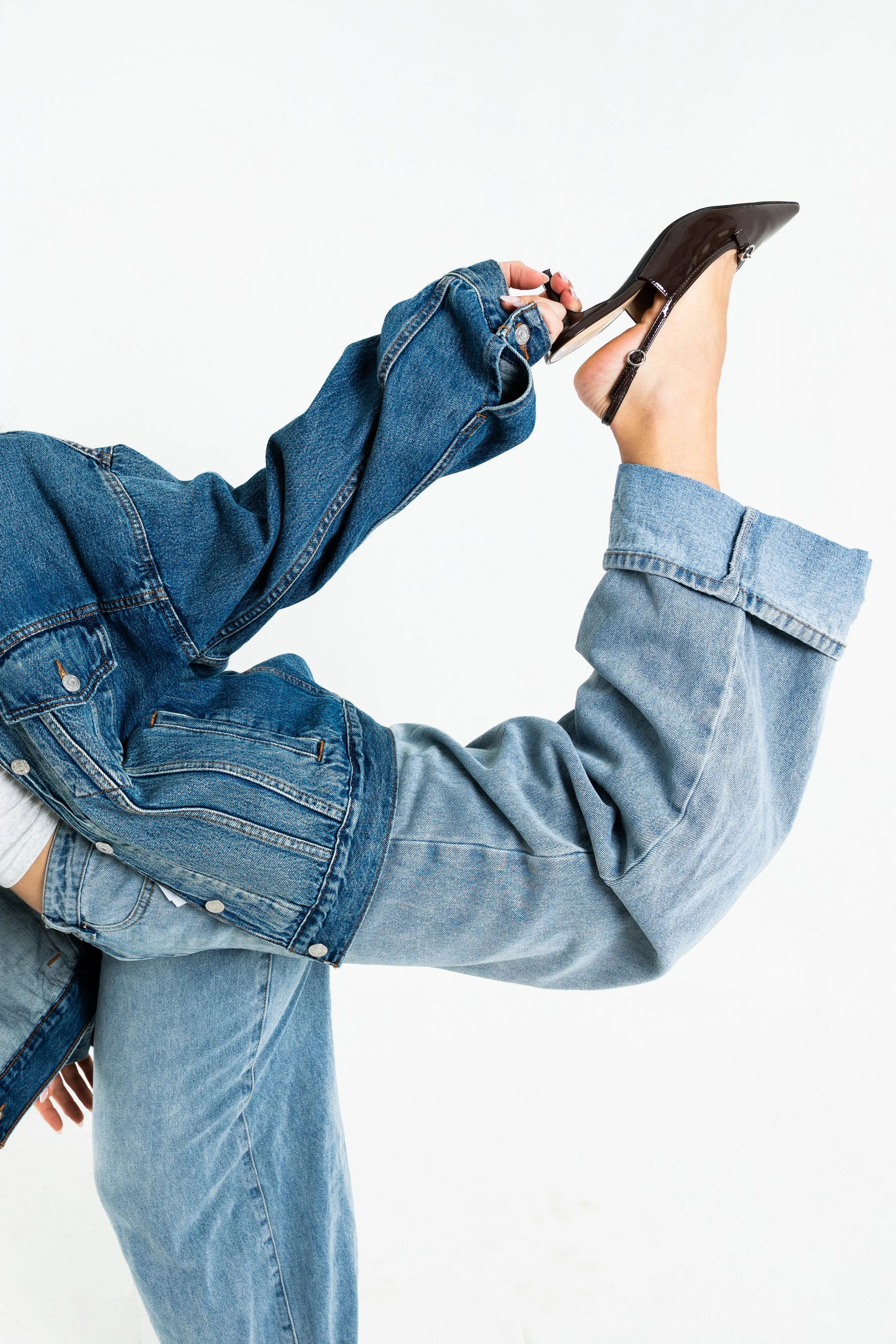 Close-up of two people in denim jeans and jackets, one holding a high-heeled shoe with straps in front of a white background.
